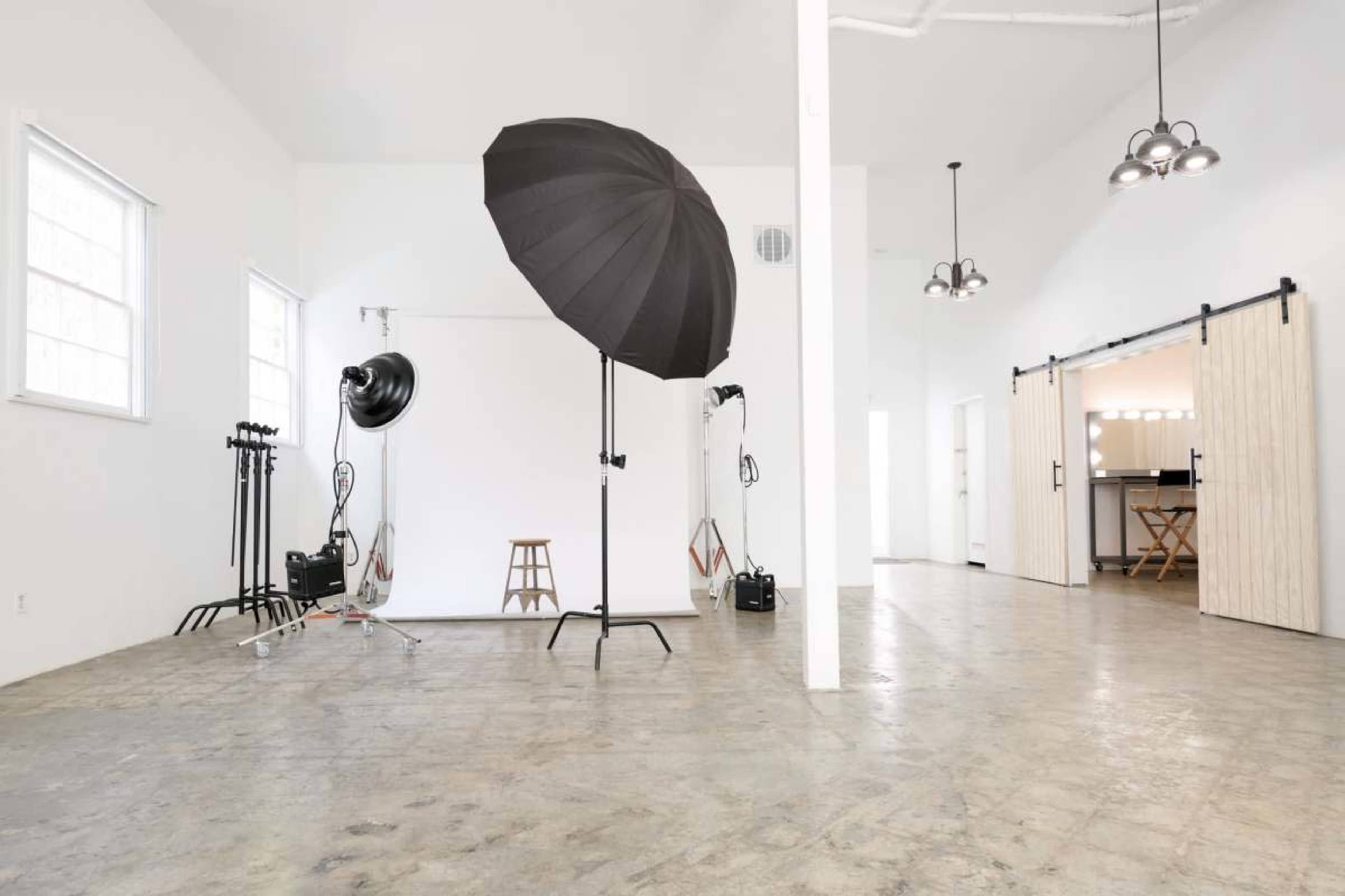 The image shows a spacious photography studio featuring a large black umbrella, light stands, a wooden stool, and a kitchen area in the background.