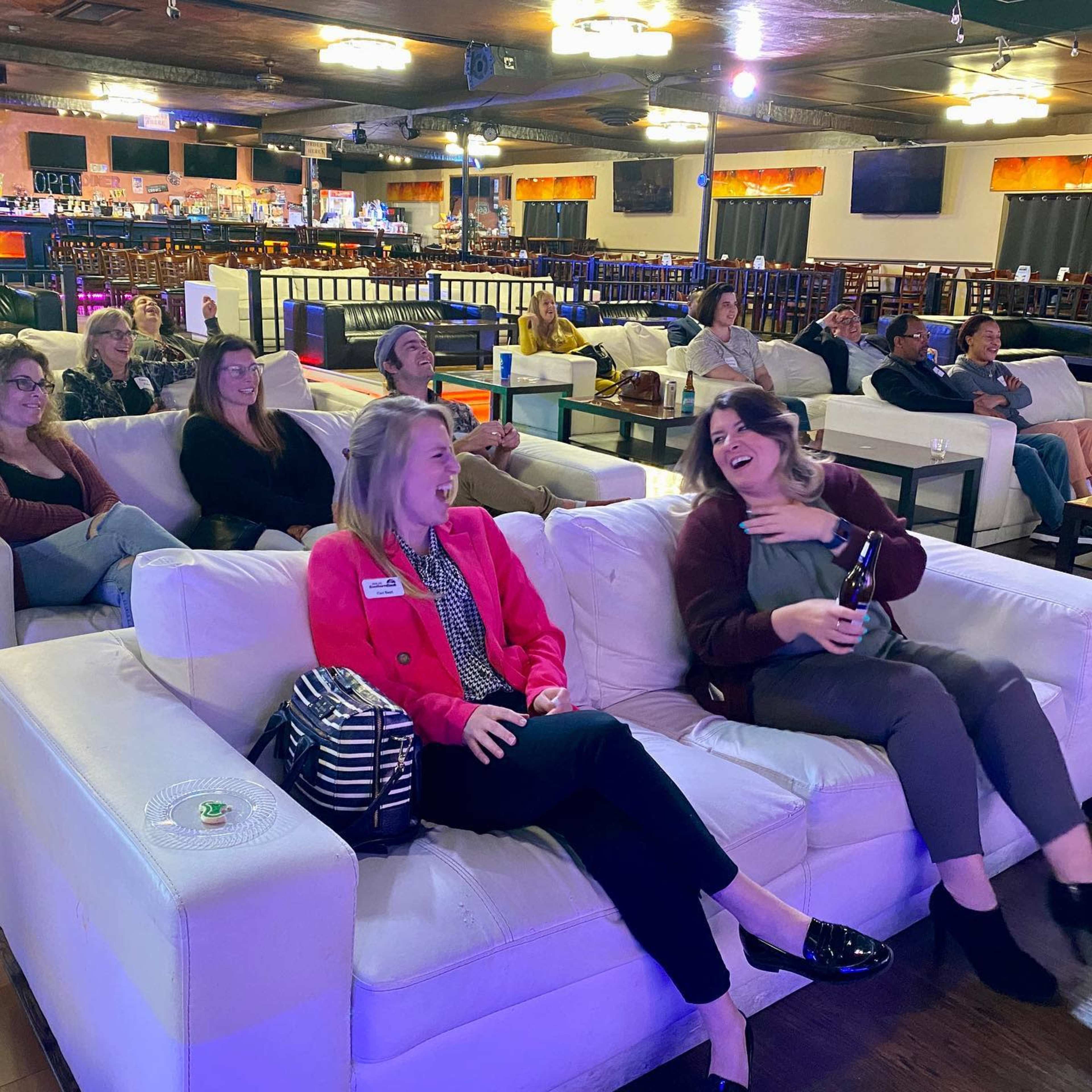 A group of people sits on white couches in a brightly lit venue, some laughing and engaging with each other.