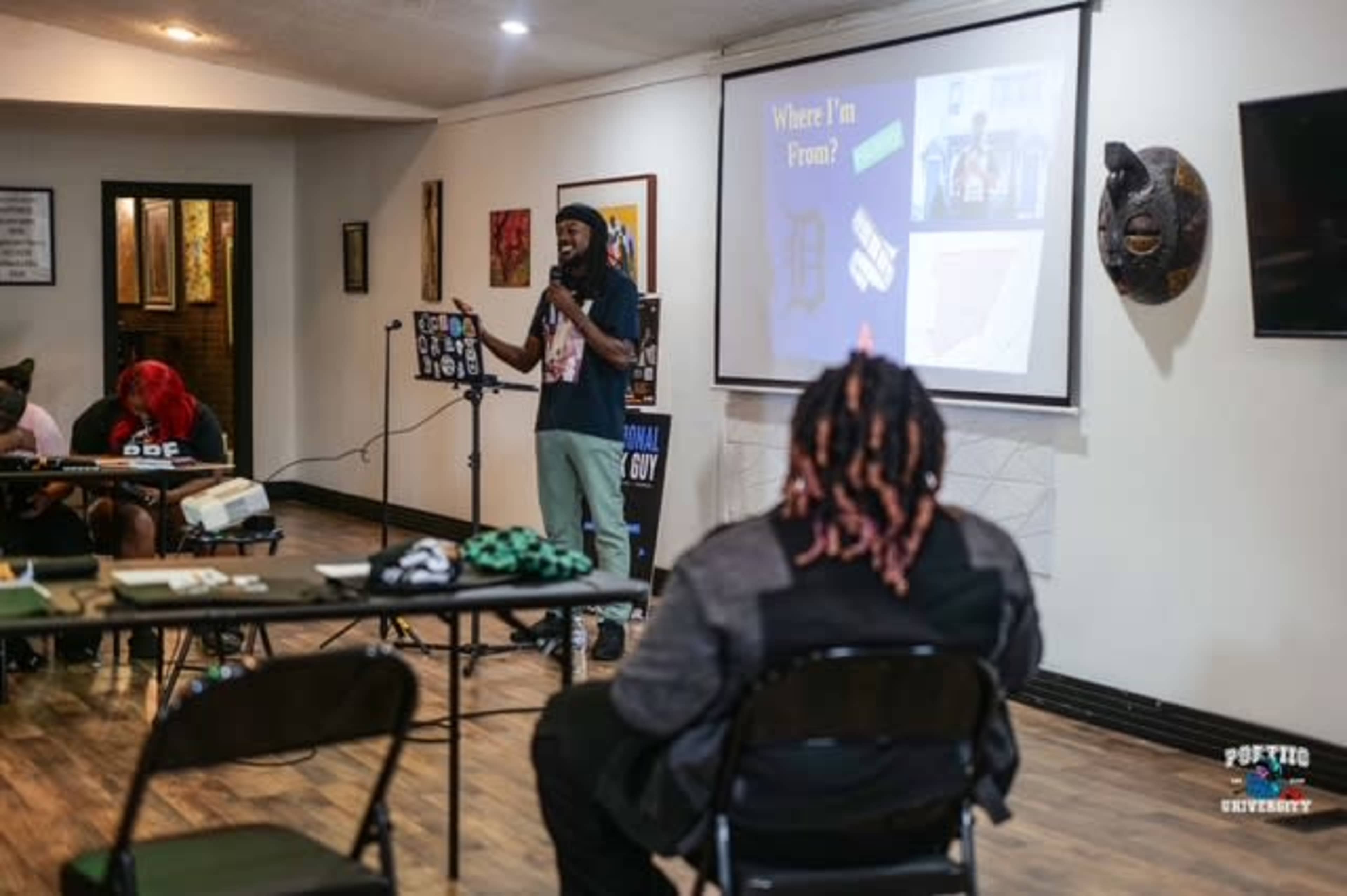 A speaker stands in front of a screen, presenting to an audience in a room decorated with artwork and cultural items.