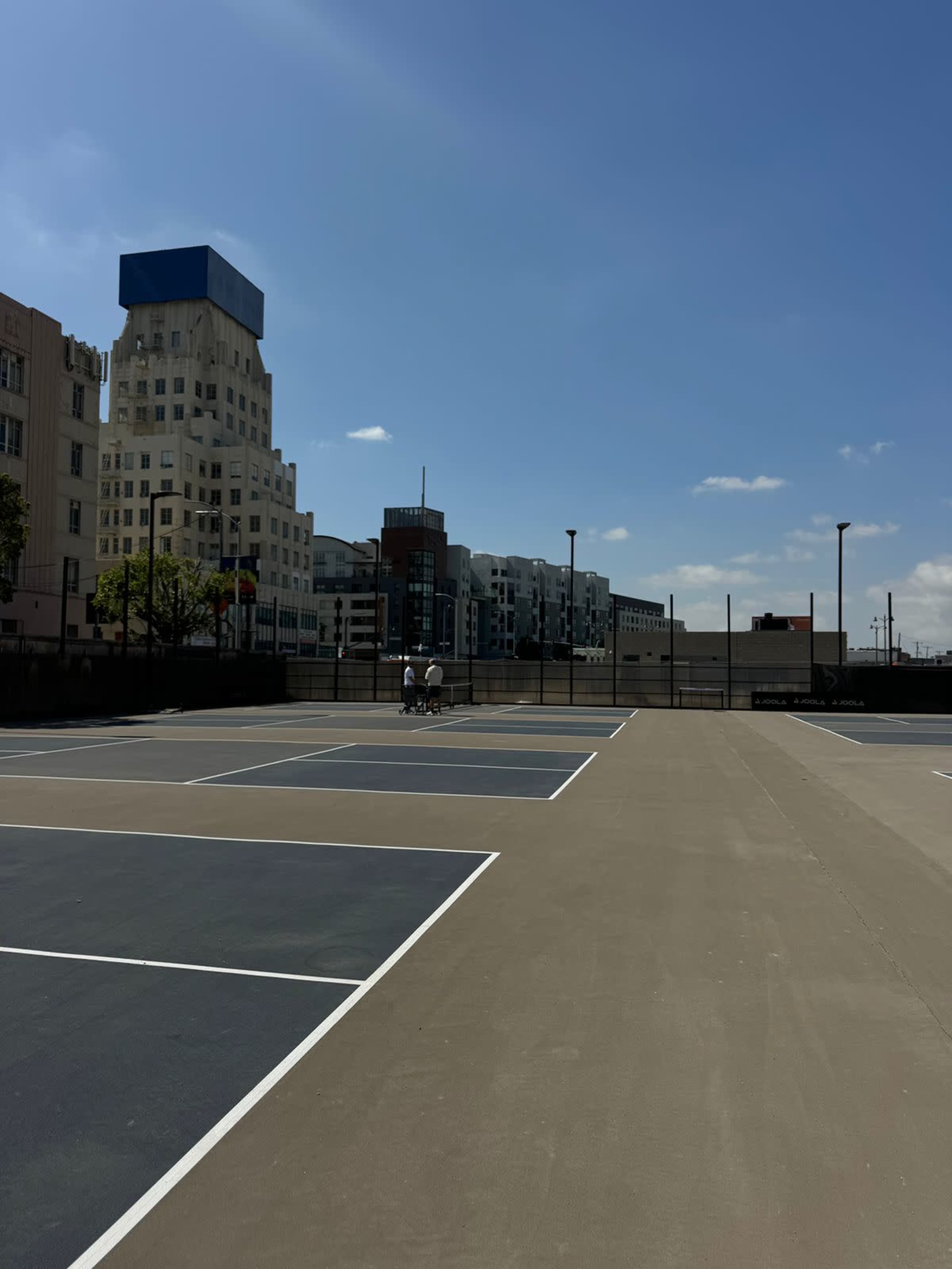 The image shows a tennis court with two players in the foreground, set against a backdrop of urban buildings under a clear blue sky.