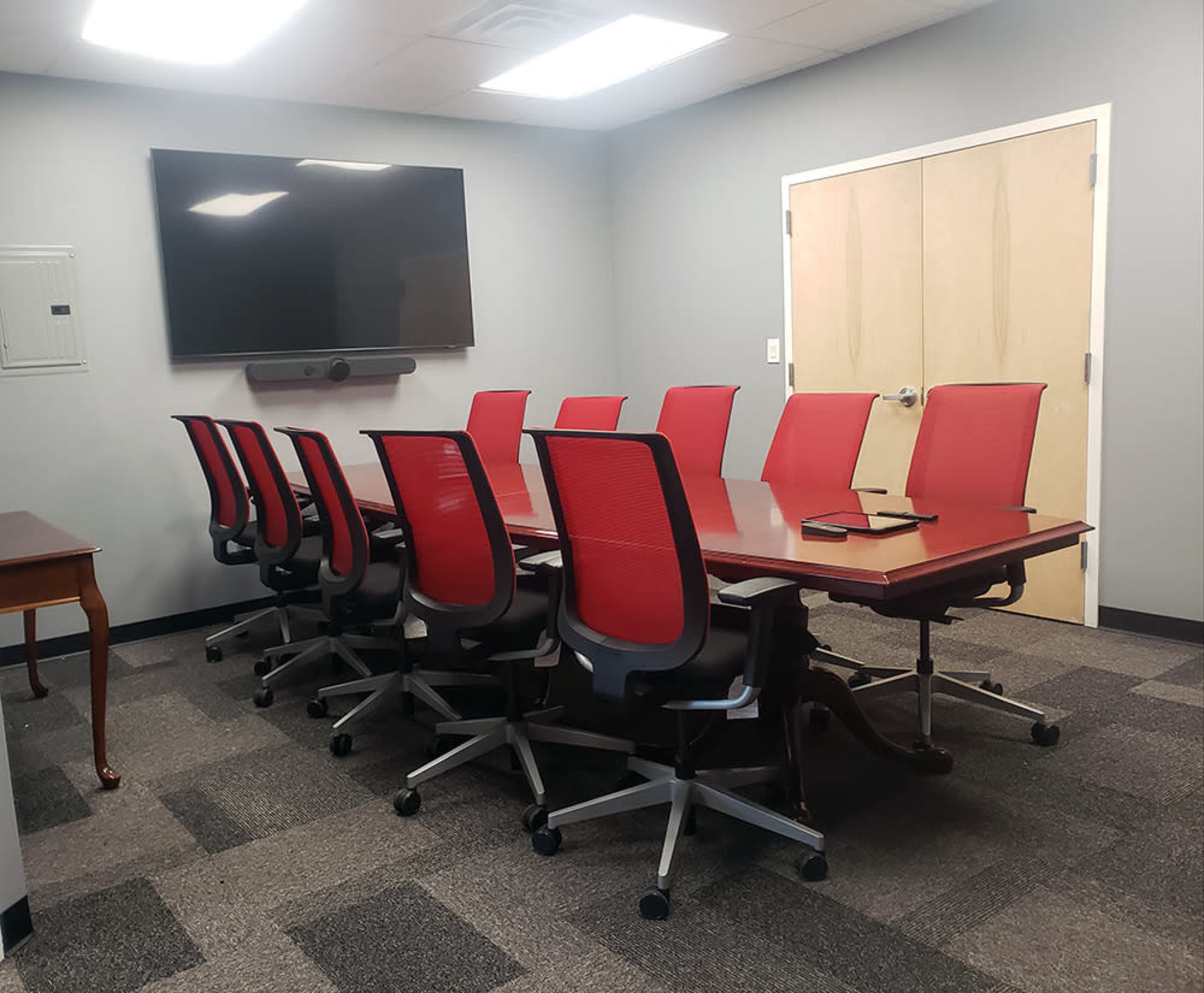 A conference room with a large rectangular table surrounded by ten red ergonomic chairs, a wall-mounted screen, and a wooden side table.