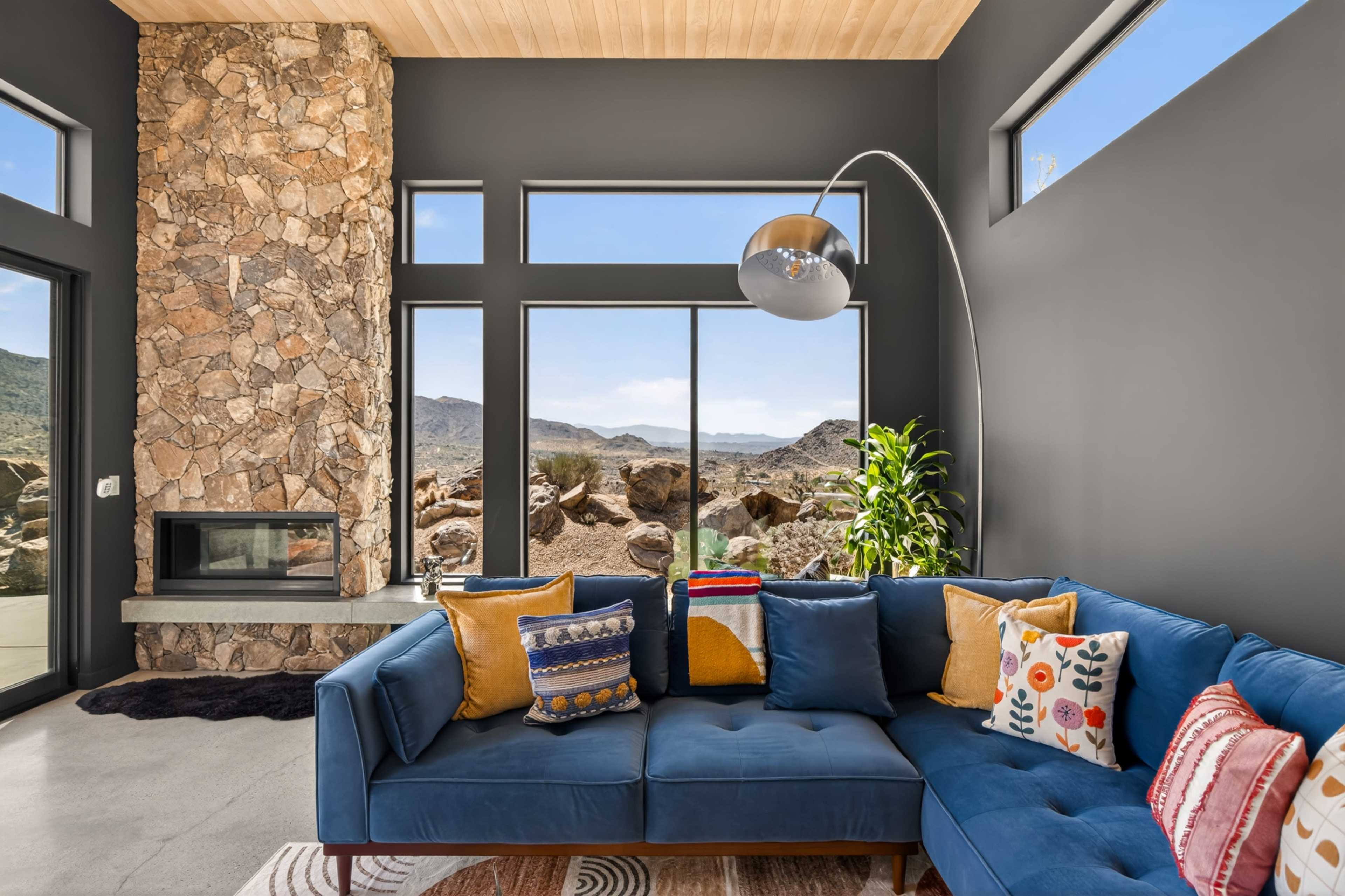 A modern living room features a blue sectional sofa adorned with colorful pillows, a stone accent wall, and large windows that showcase a mountainous landscape.
