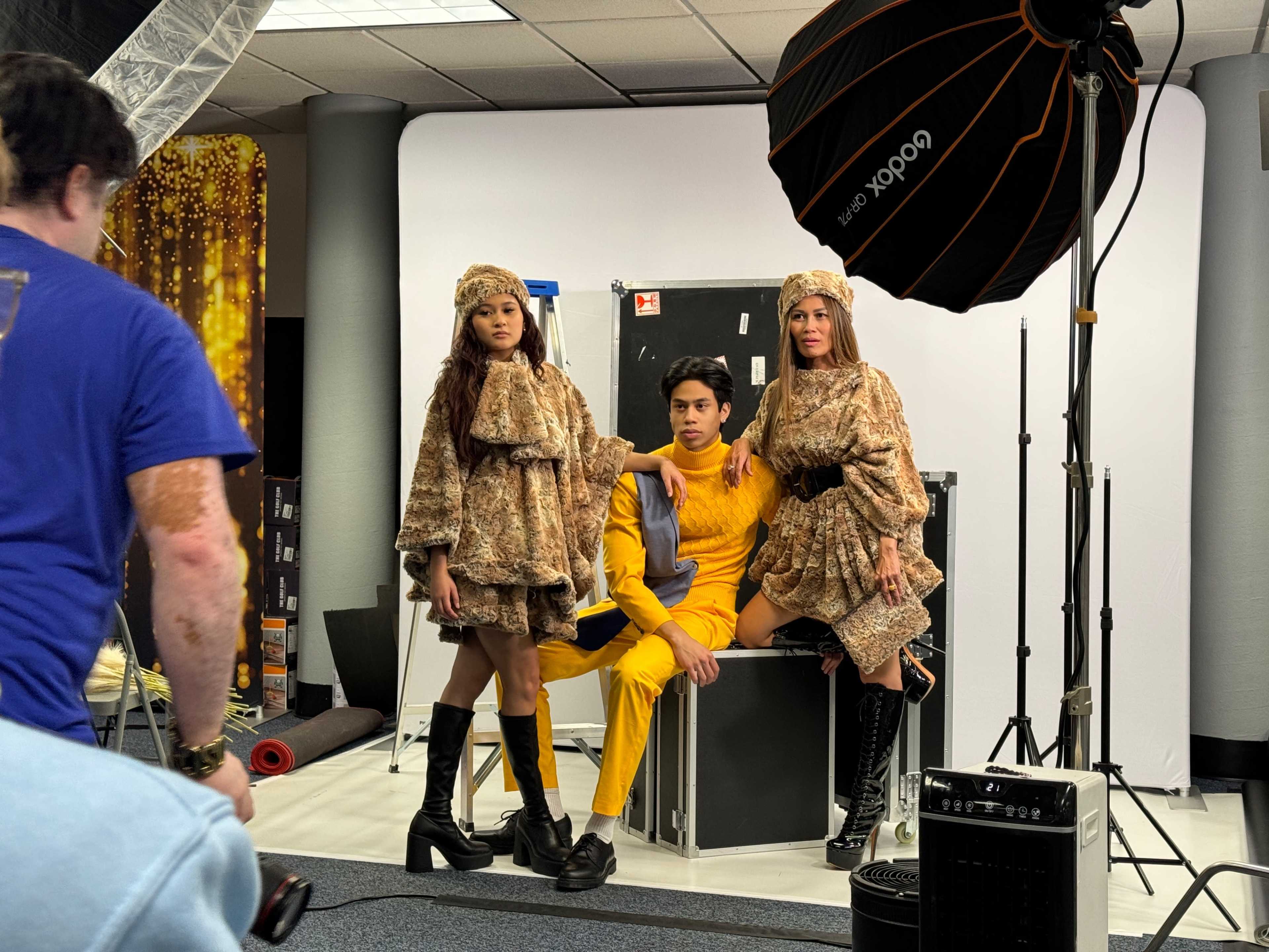 Two women in designer outfits pose alongside a man in a yellow suit during a photoshoot in a studio setting.