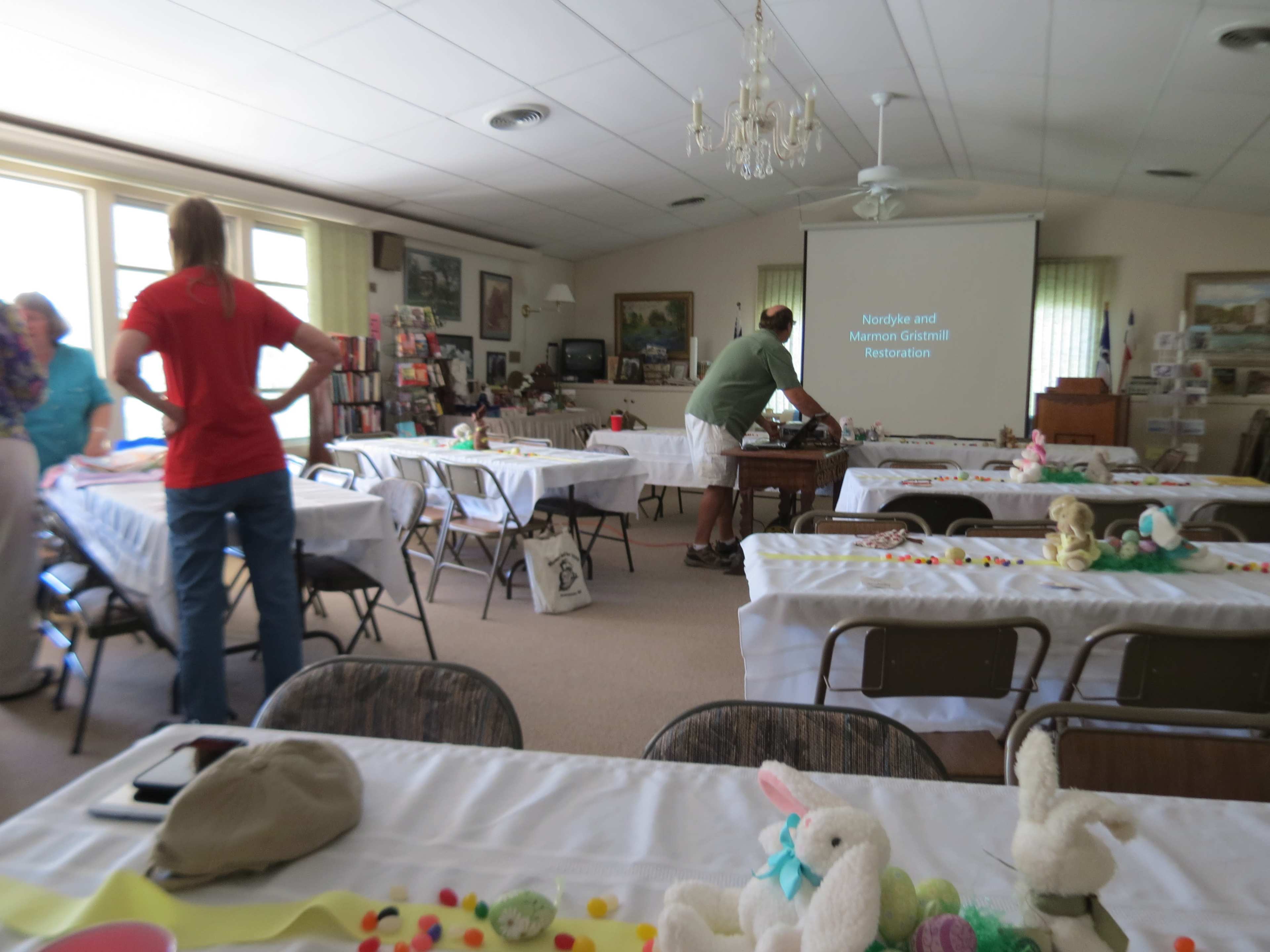 A group of people is gathered in a community room with tables set up for a crafting event, featuring various supplies and a presentation screen displaying "Norfolk and Manson Grant Restoration."