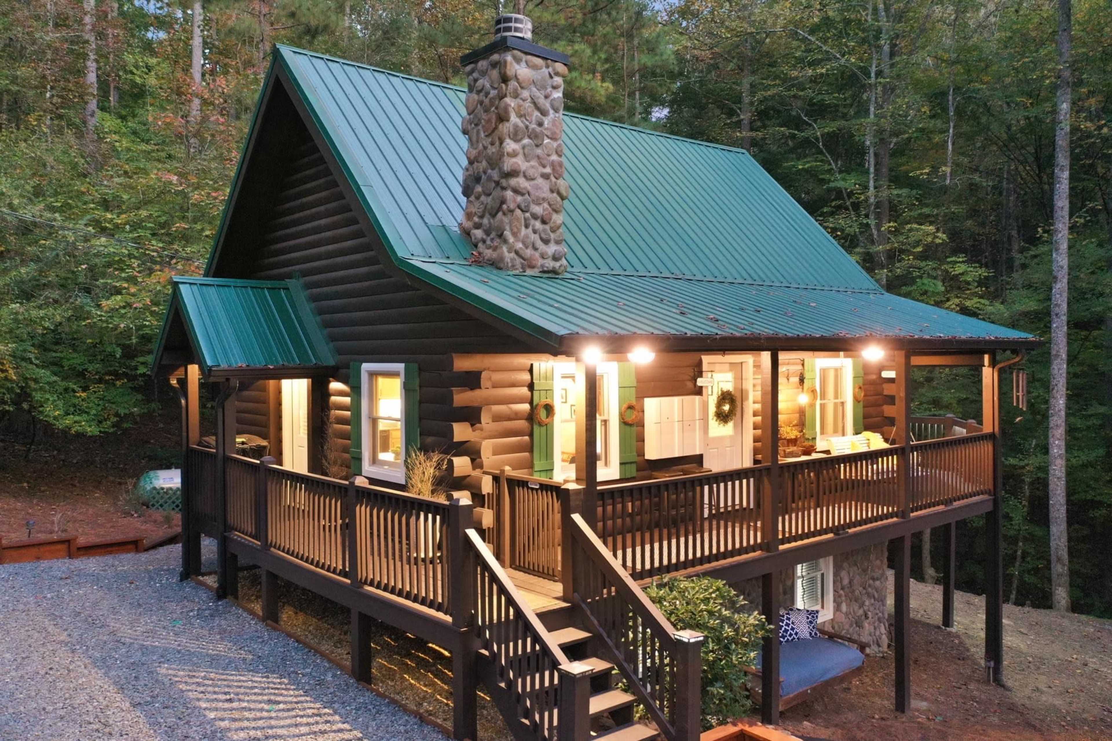 A log cabin with a green metal roof features a stone chimney and a spacious porch surrounded by trees.
