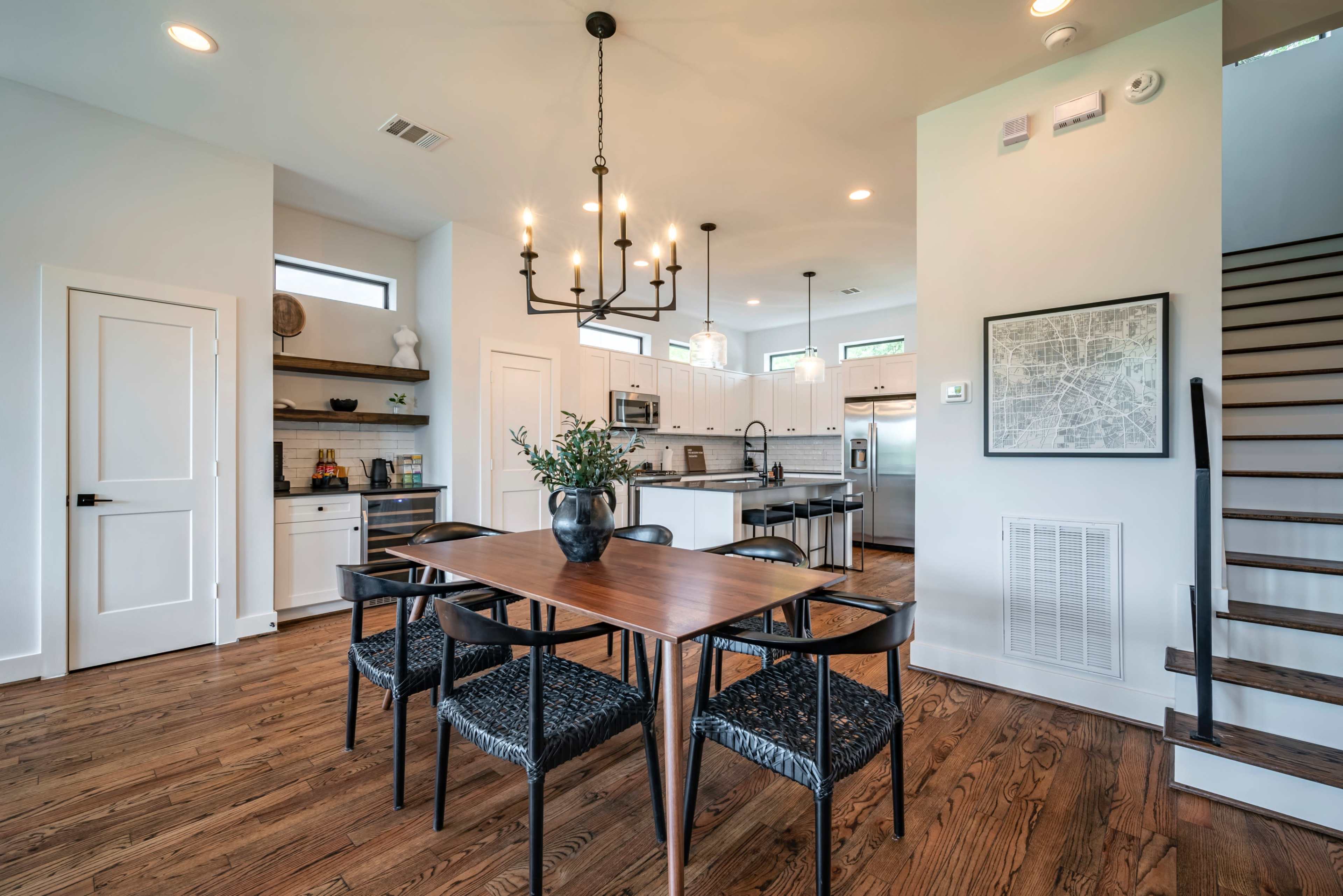 A dining area features a wooden table surrounded by black chairs, with a modern kitchen and staircase visible in the background.