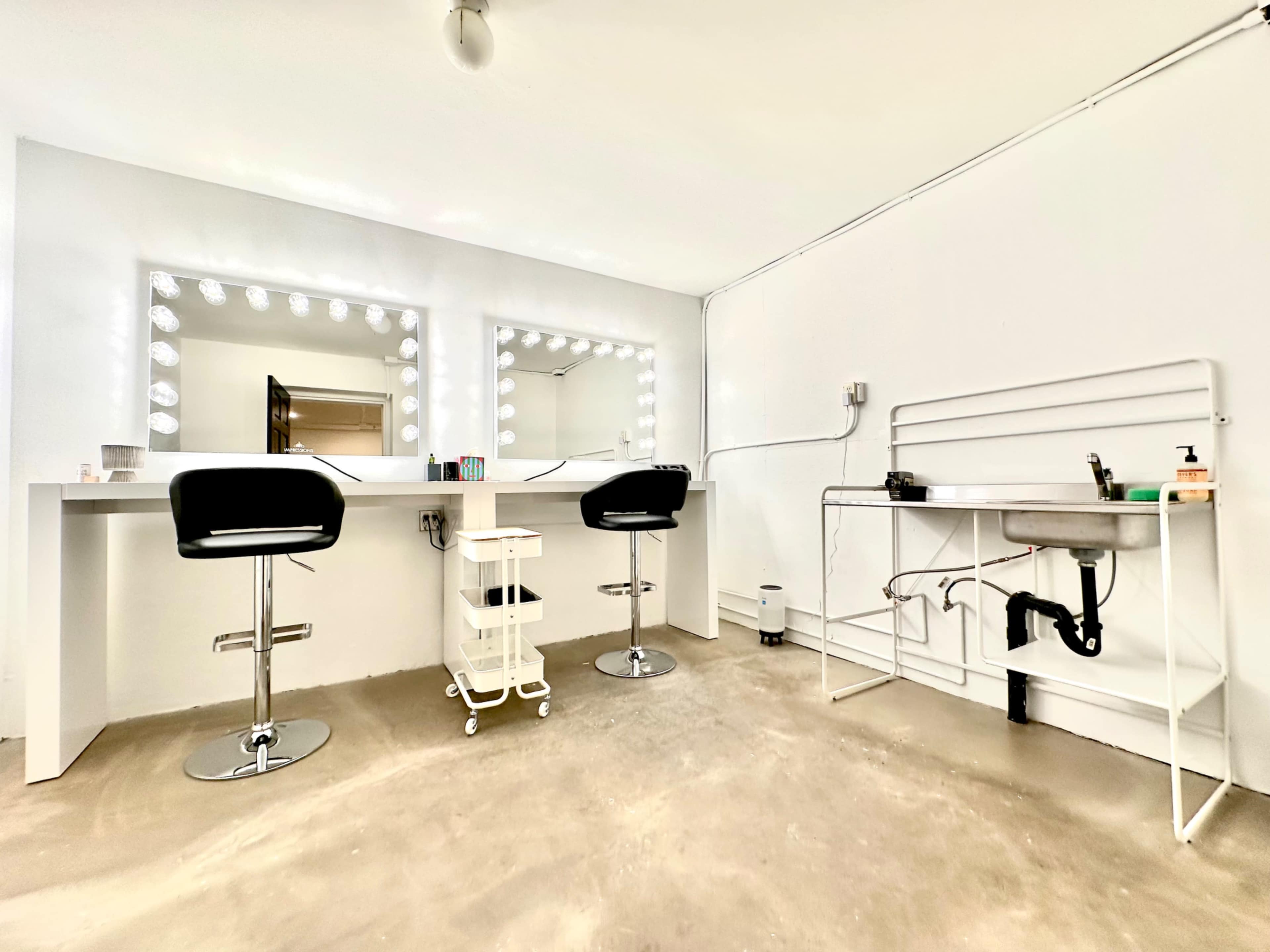 The image shows a minimalist beauty salon interior with two illuminated mirrors above black bar stools and a sink area beside a metal cart.