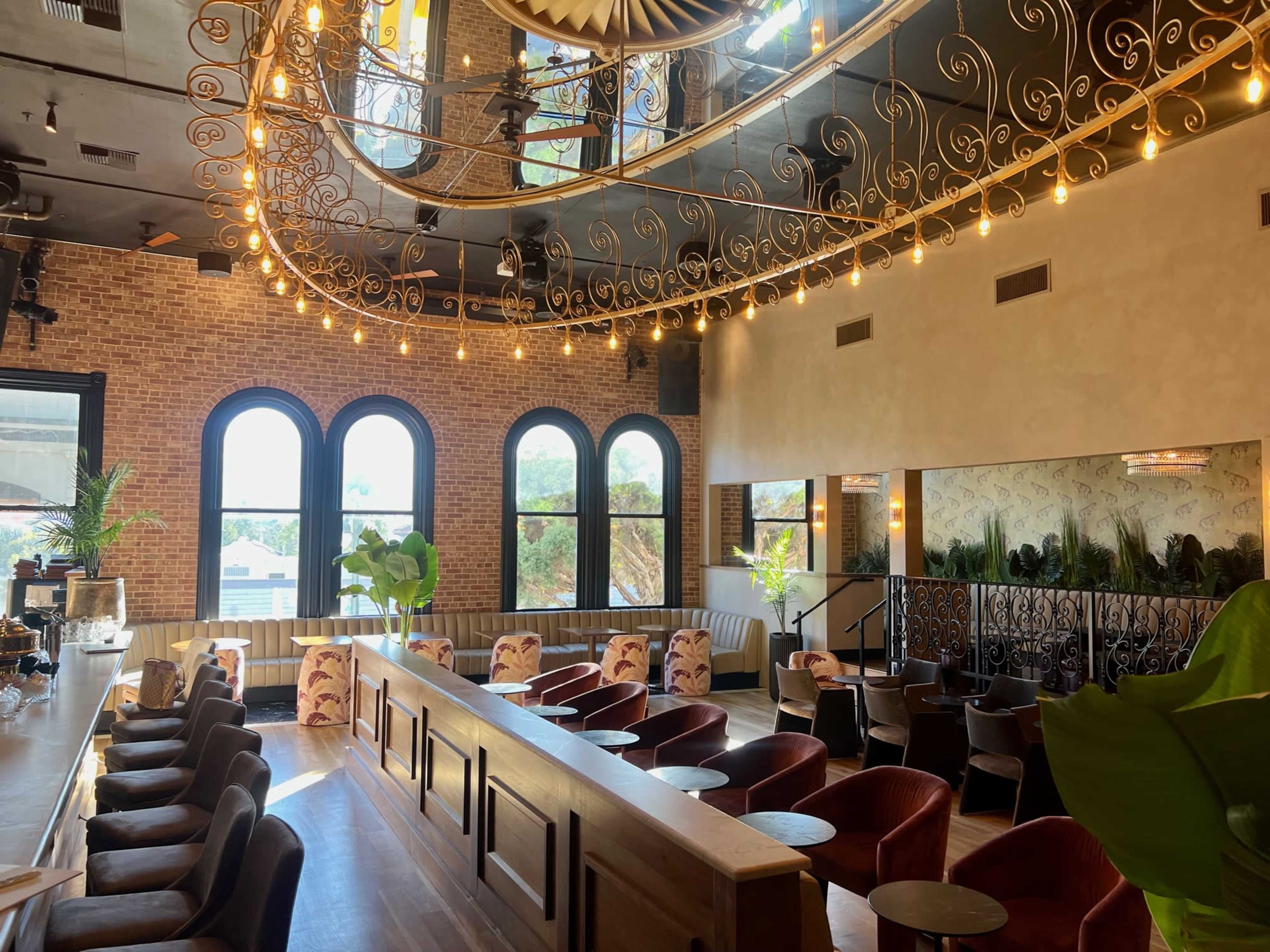 The image shows a stylish restaurant interior featuring arched windows, a high ceiling adorned with decorative light fixtures, and a mixture of tables and plush seating arrangements.