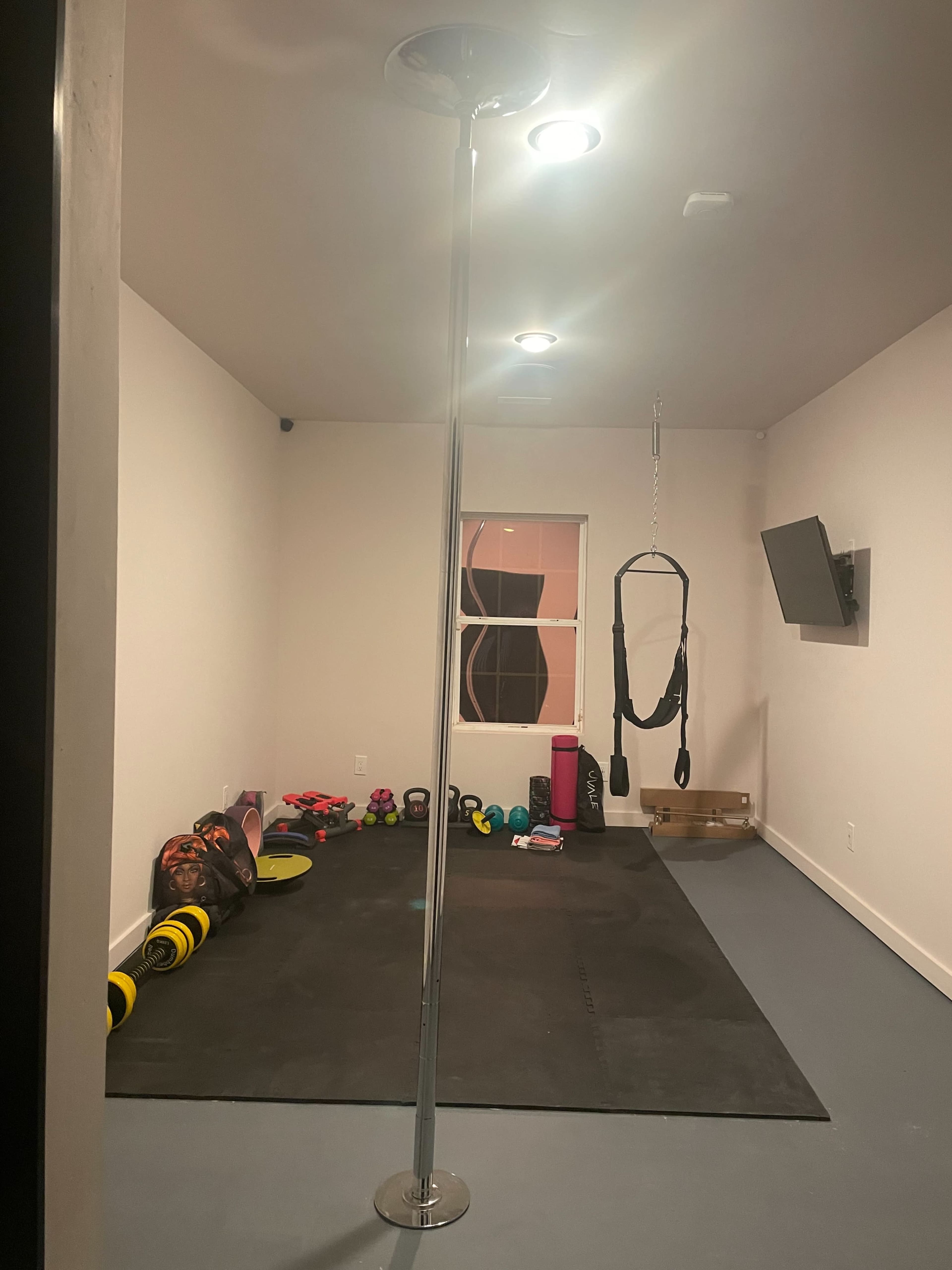A home gym with a pole in the center, weights and exercise equipment scattered on a black mat, and a television mounted on the wall.