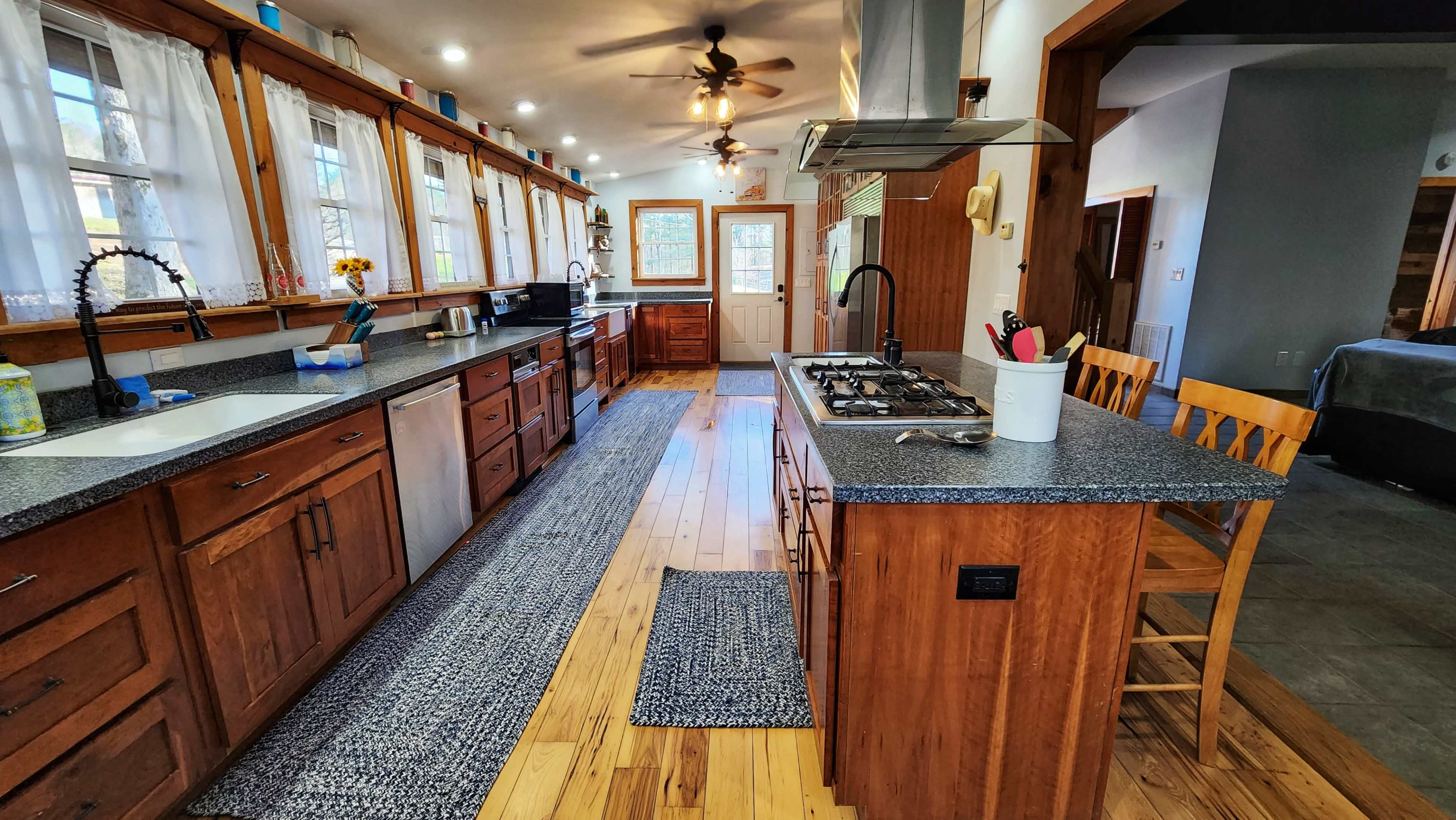 A modern kitchen with wooden cabinets, a central island featuring a gas stove, and large windows allowing natural light to enter the space.