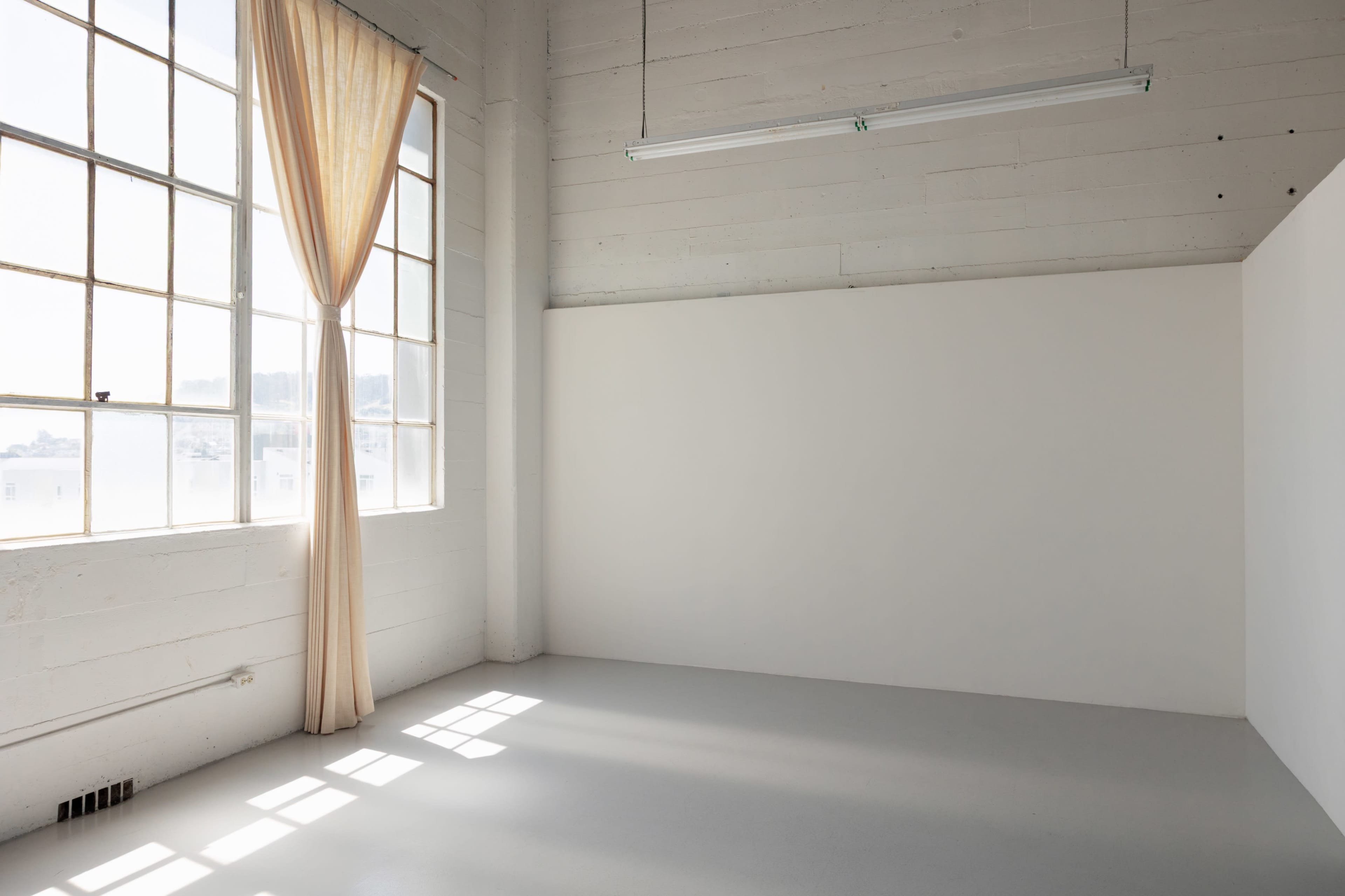 The image shows an empty room with large windows letting in natural light and a single curtain hanging beside an unadorned white wall.