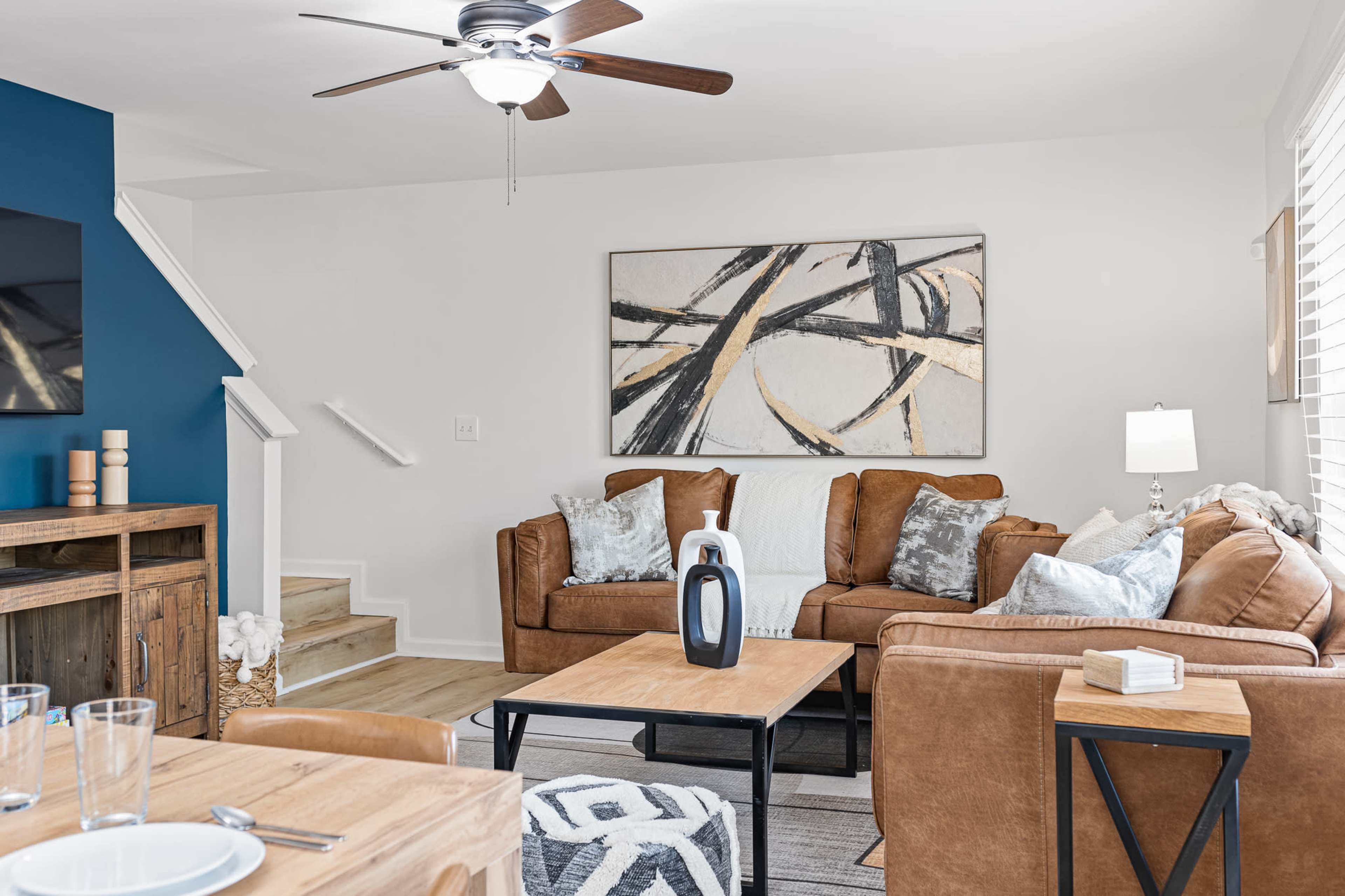 A modern living room features a brown leather sectional sofa, a wooden coffee table, and a blue accent wall, with a staircase visible in the background.