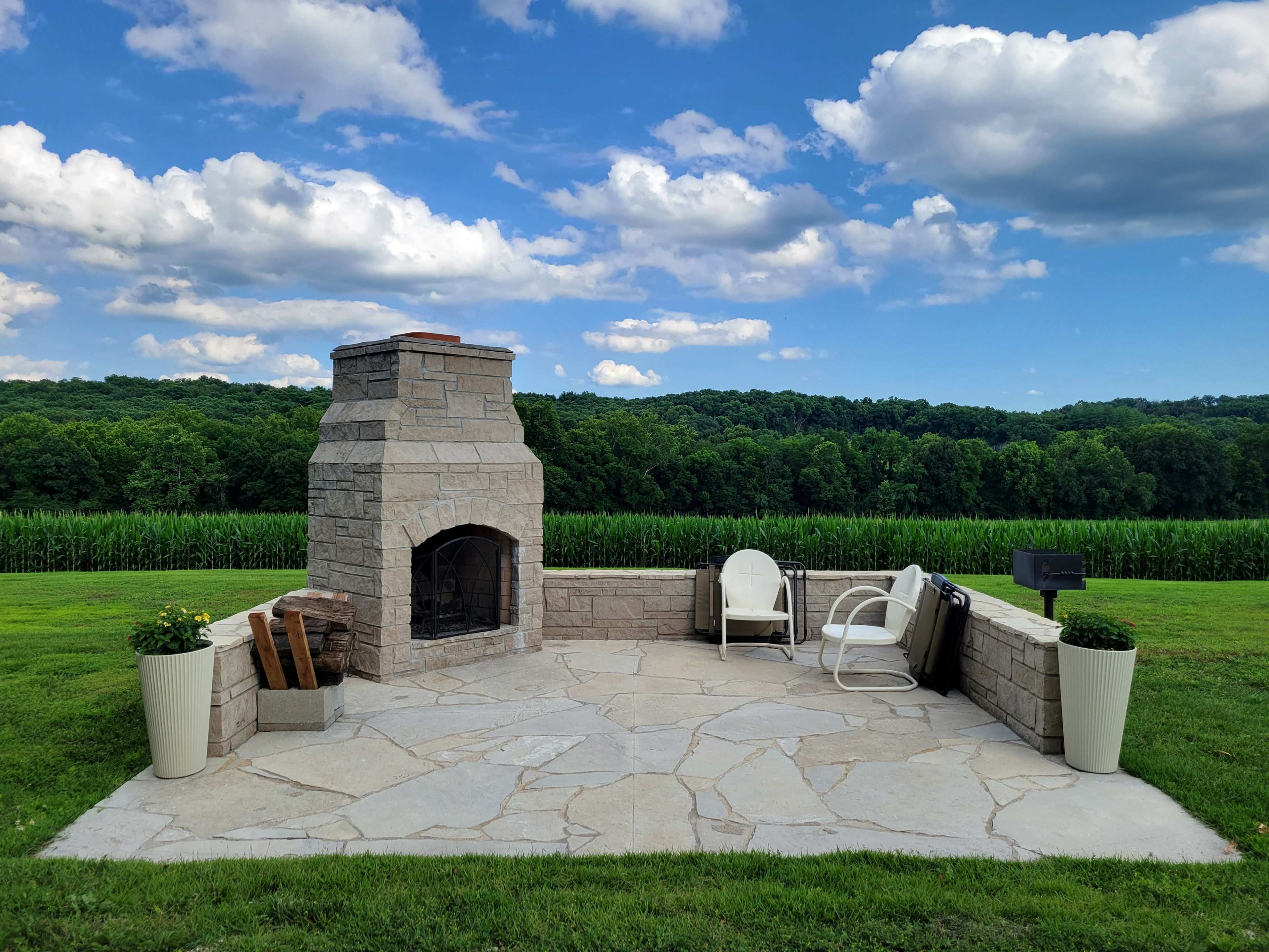 The image shows a stone outdoor fireplace on a patio, surrounded by green grass and hills, under a partly cloudy blue sky.