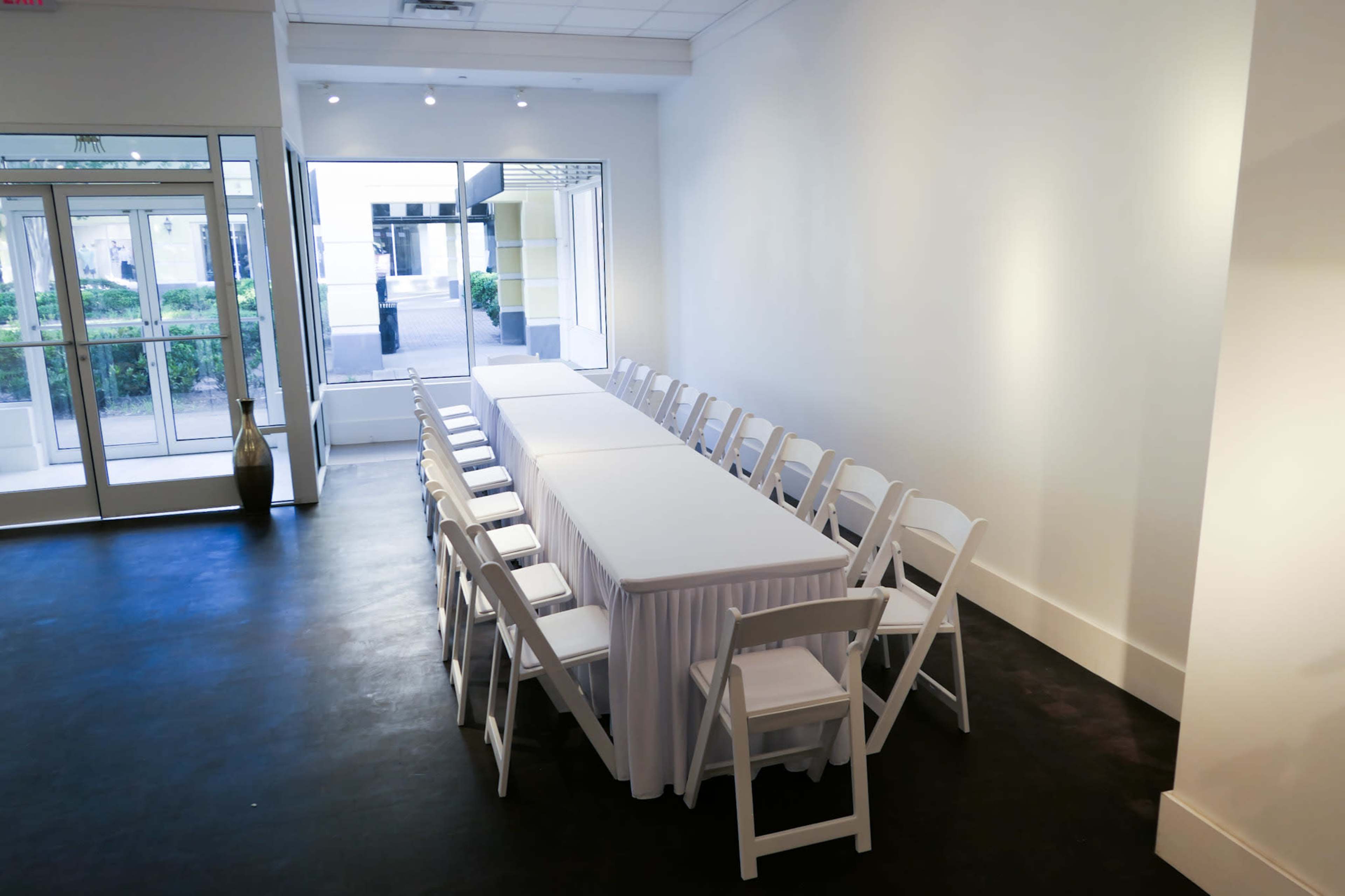 A long, rectangular table covered with a white tablecloth is set up in a bright, minimalist conference room with white walls and chairs arranged on either side.