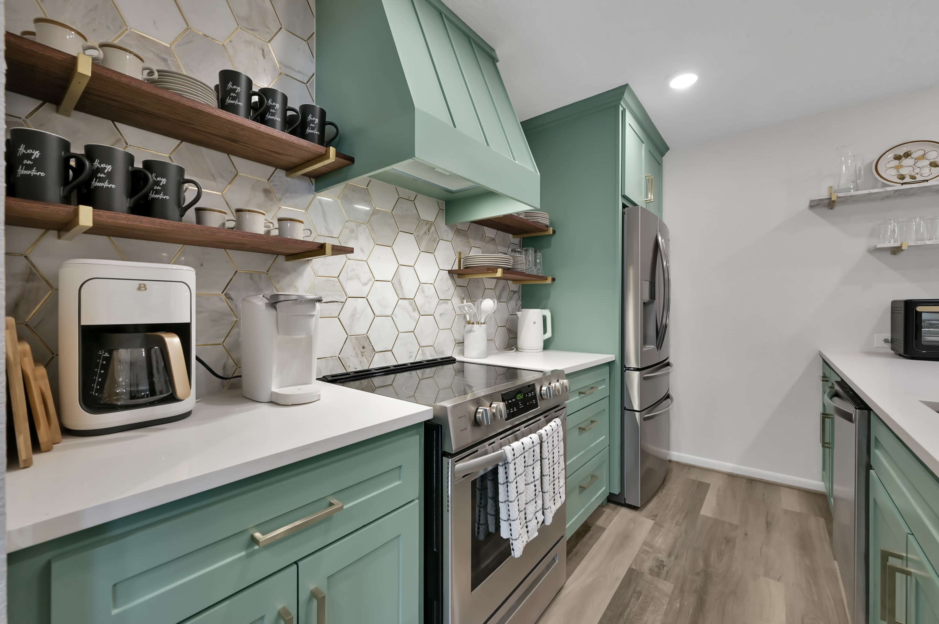 A modern kitchen features mint green cabinets, hexagonal tile backsplash, stainless steel appliances, and floating shelves with black mugs.