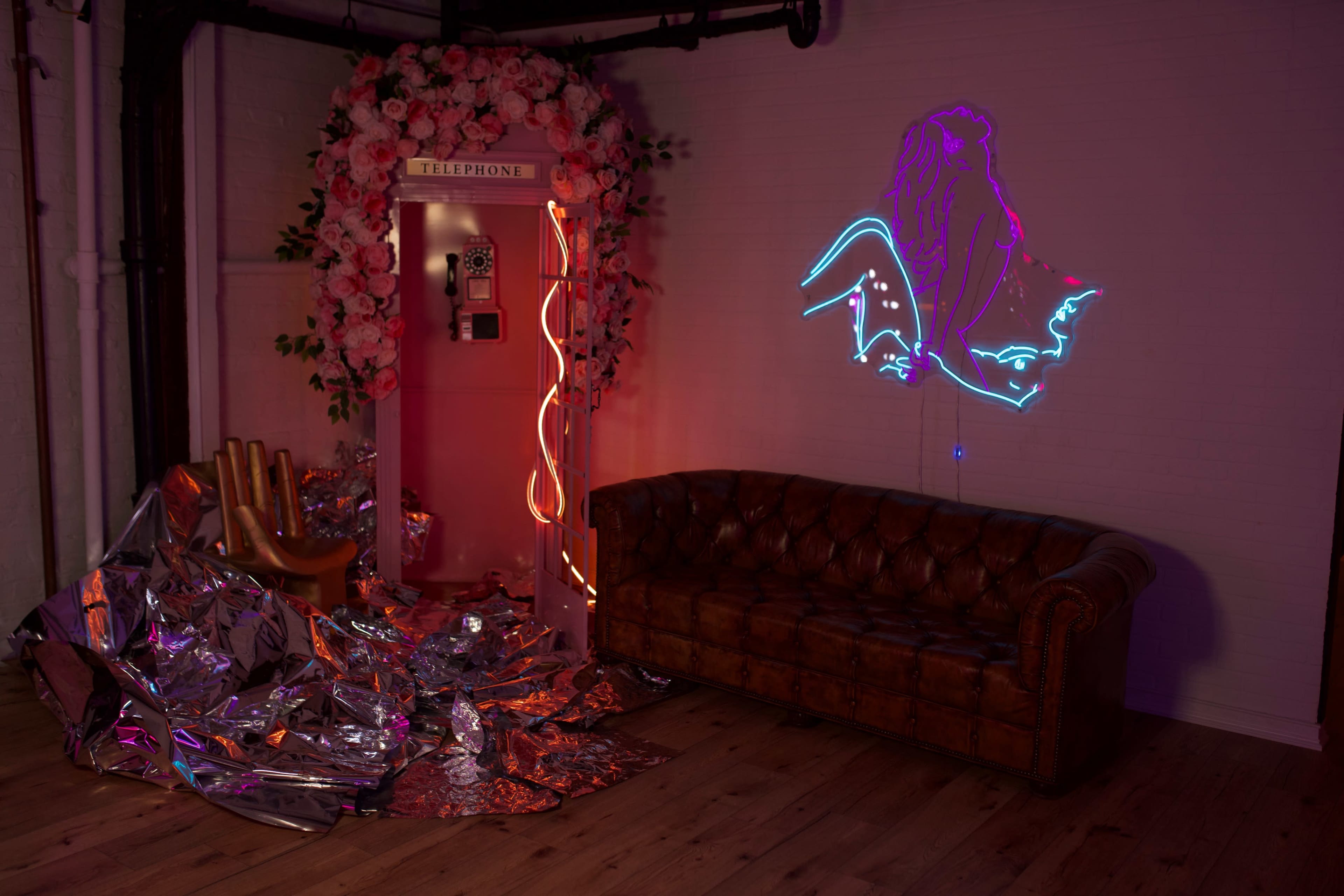 A vintage telephone booth surrounded by pink flowers, a neon lion graphic on the wall, a shiny silver material spread across the floor, and a brown leather sofa.