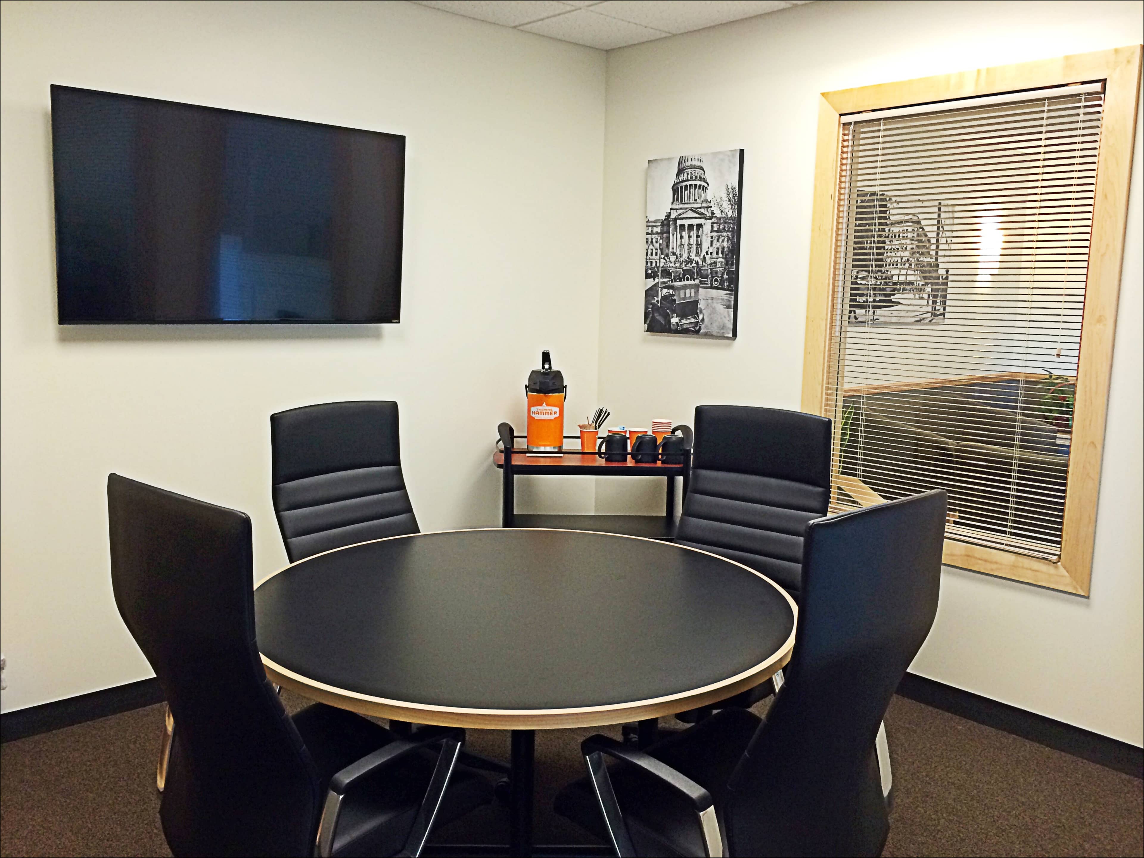 A modern conference room features a round black table surrounded by four high-backed chairs, with a wall-mounted TV and a framed black-and-white photo in the background.