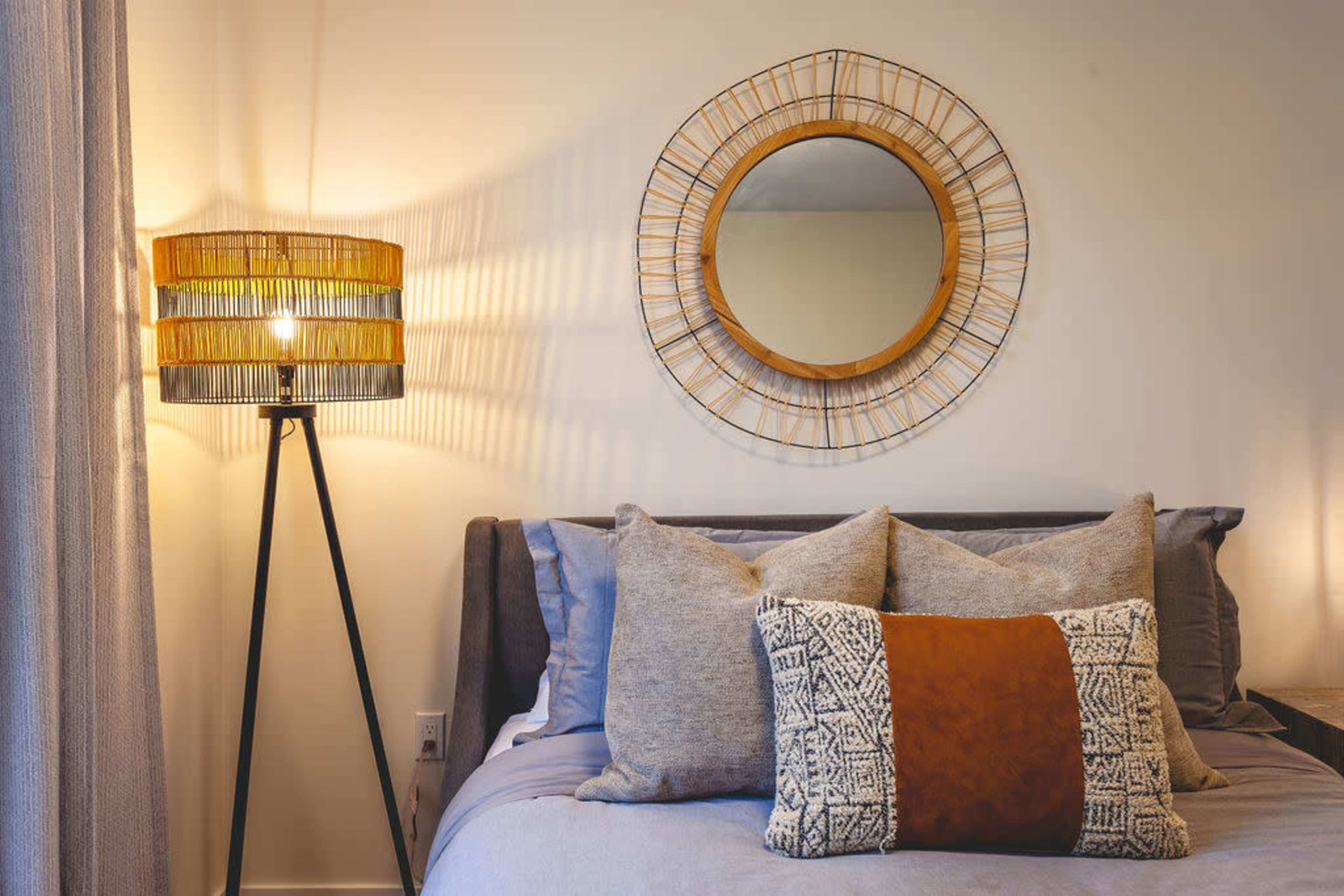 The image shows a cozy bedroom featuring a bed with decorative pillows, a round mirror on the wall, and a floor lamp with a wicker shade.