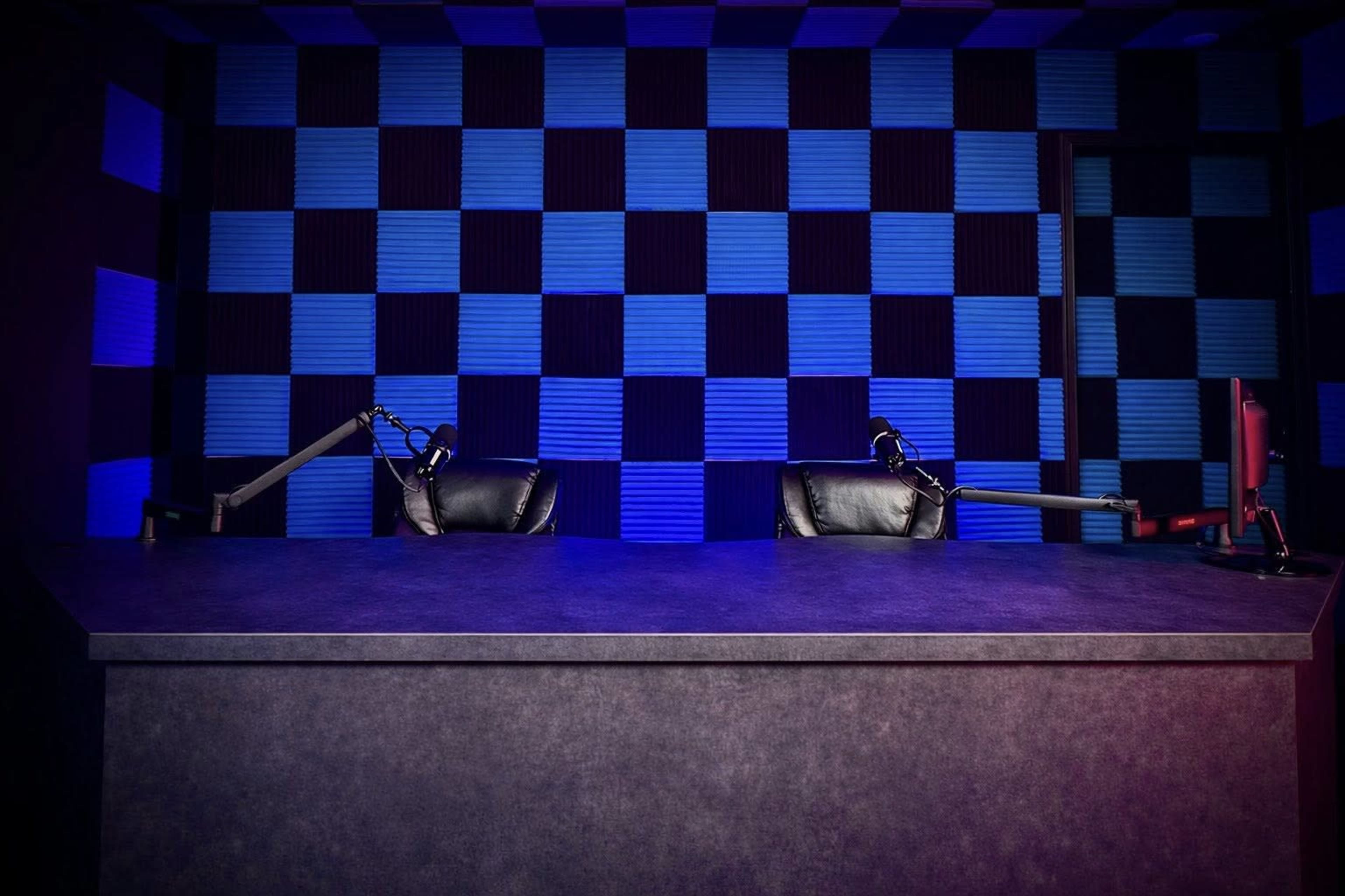 The image shows a podcast studio setup featuring a dark gray table with two black chairs and blue and black acoustic panels on the wall behind it.