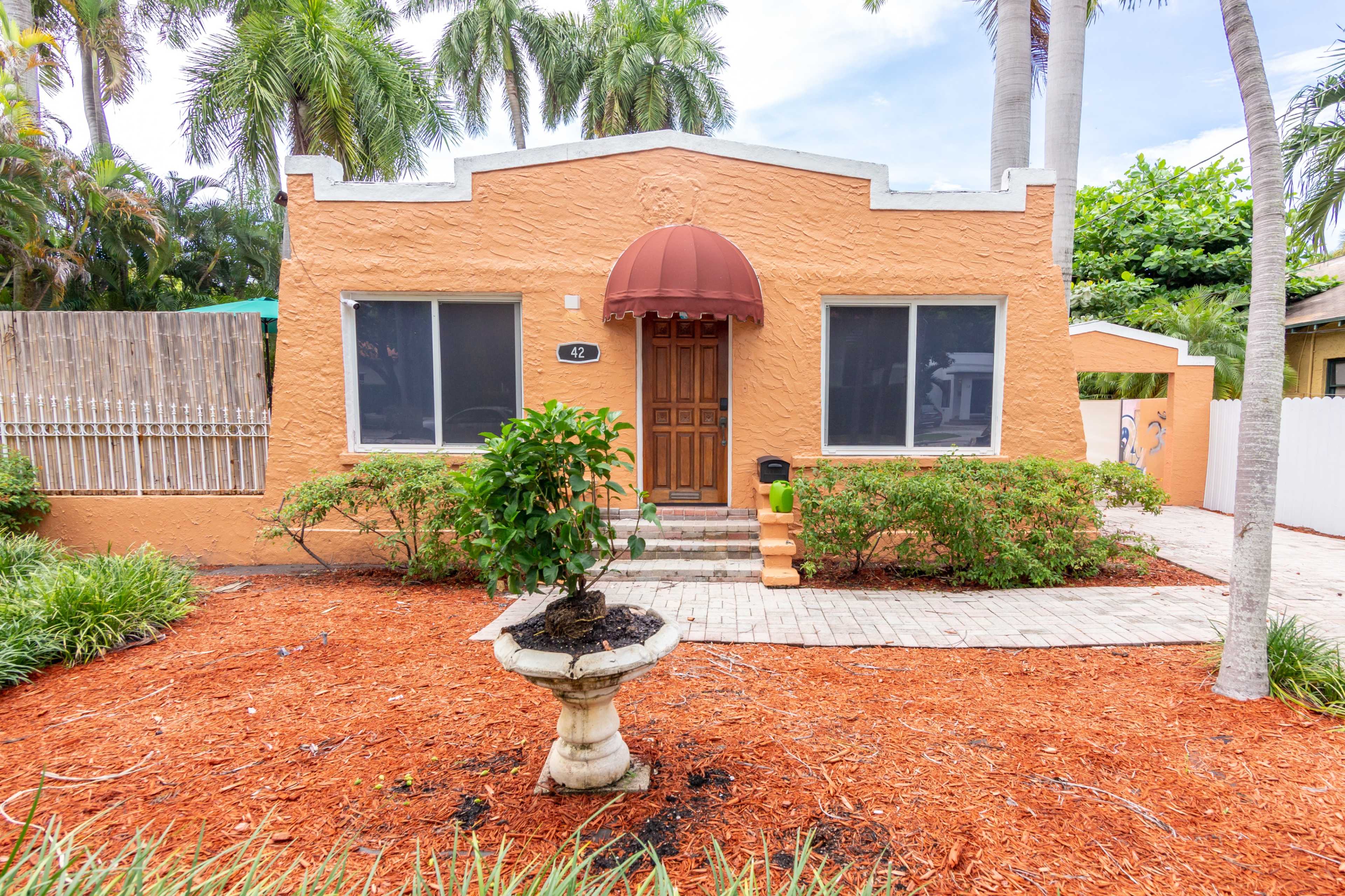A single-story orange house with a red awning over the front door, surrounded by greenery and a pathway of bricks and mulch.