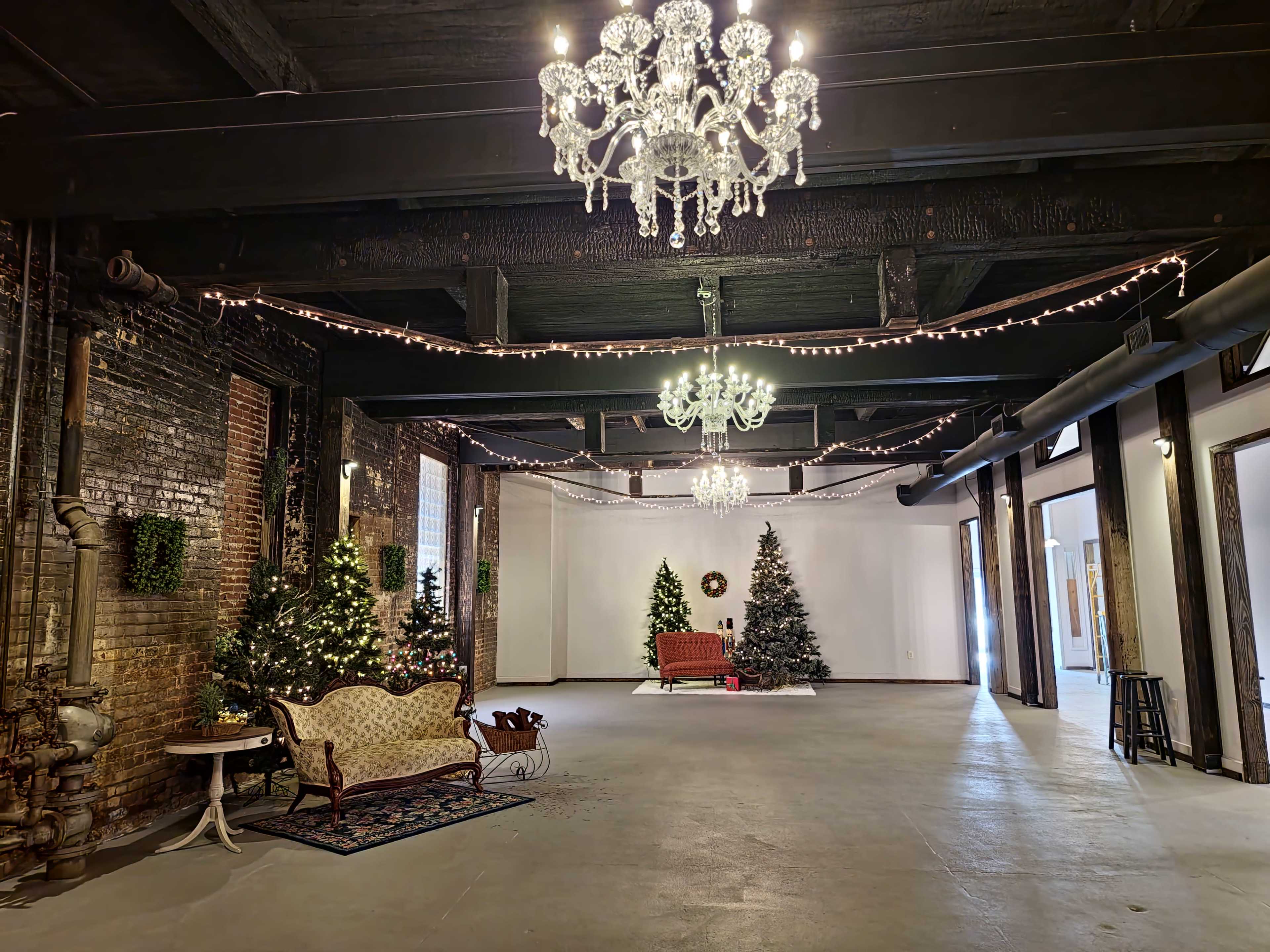 An expansive indoor space decorated for the holidays, featuring several Christmas trees, elegant chandeliers, and soft lighting.