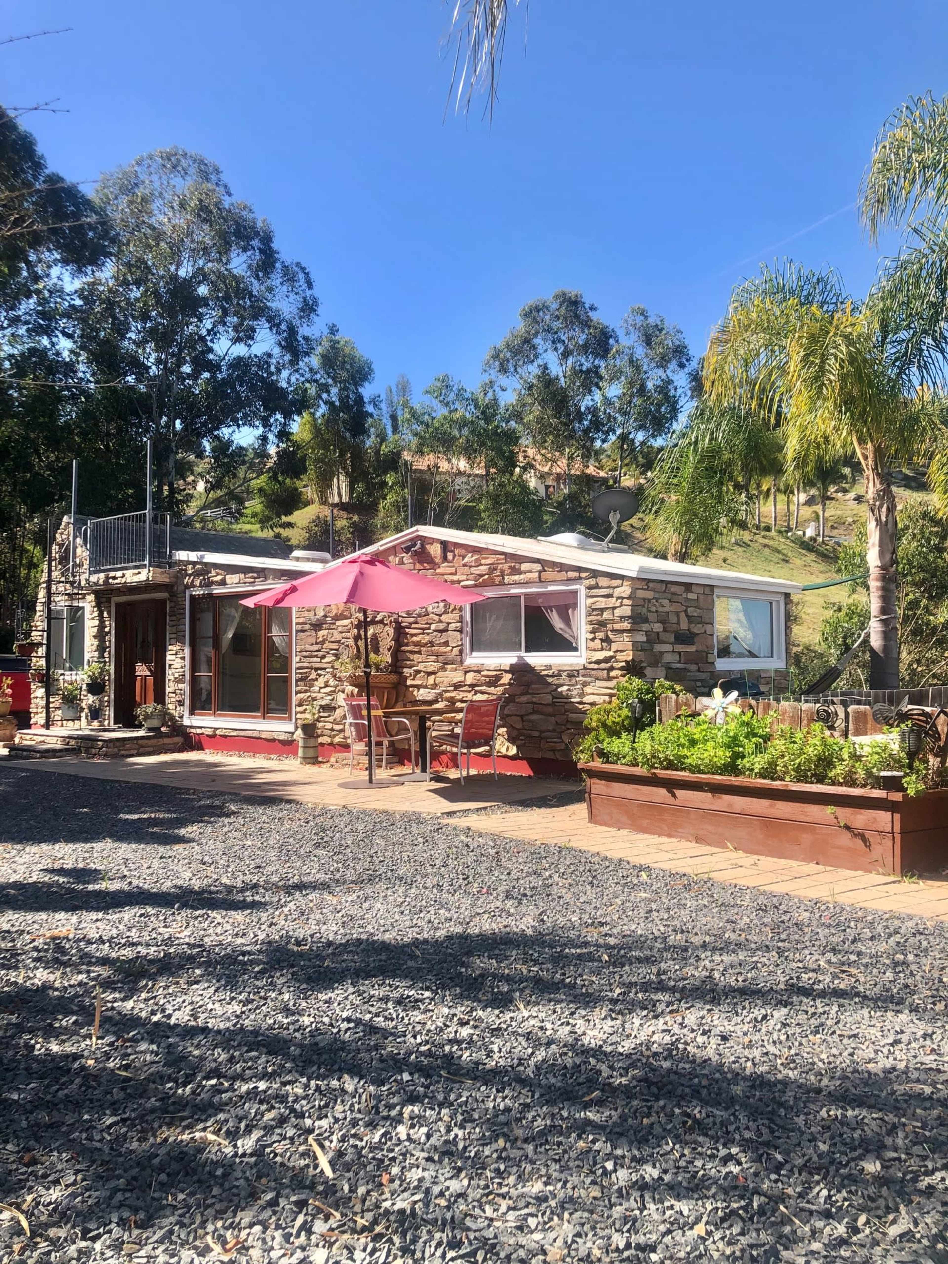 A small stone cottage with a pink umbrella and a garden bed is situated on a gravel area surrounded by trees and hills.