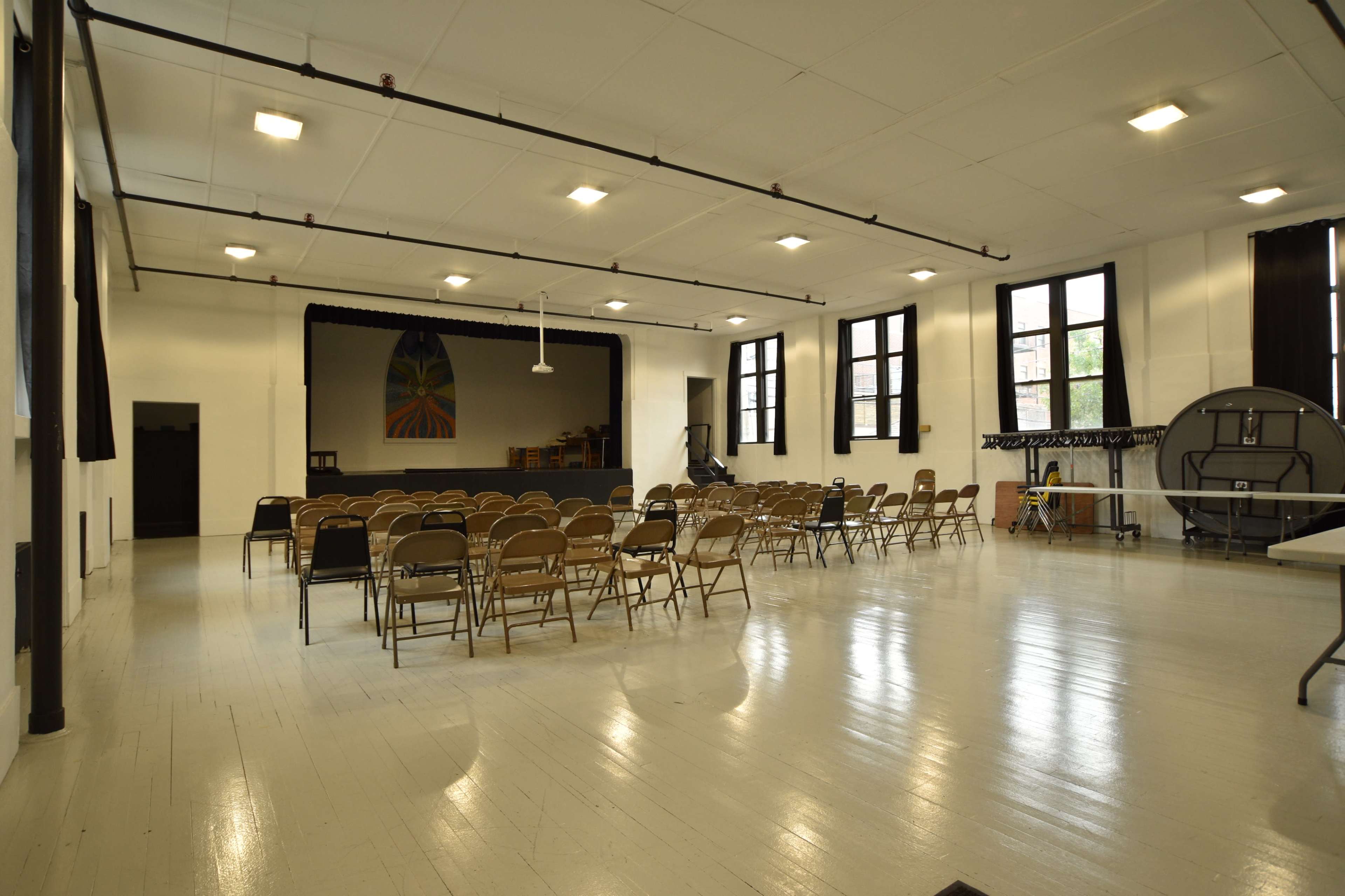 An empty auditorium with rows of folding chairs facing a stage and large windows letting in natural light.