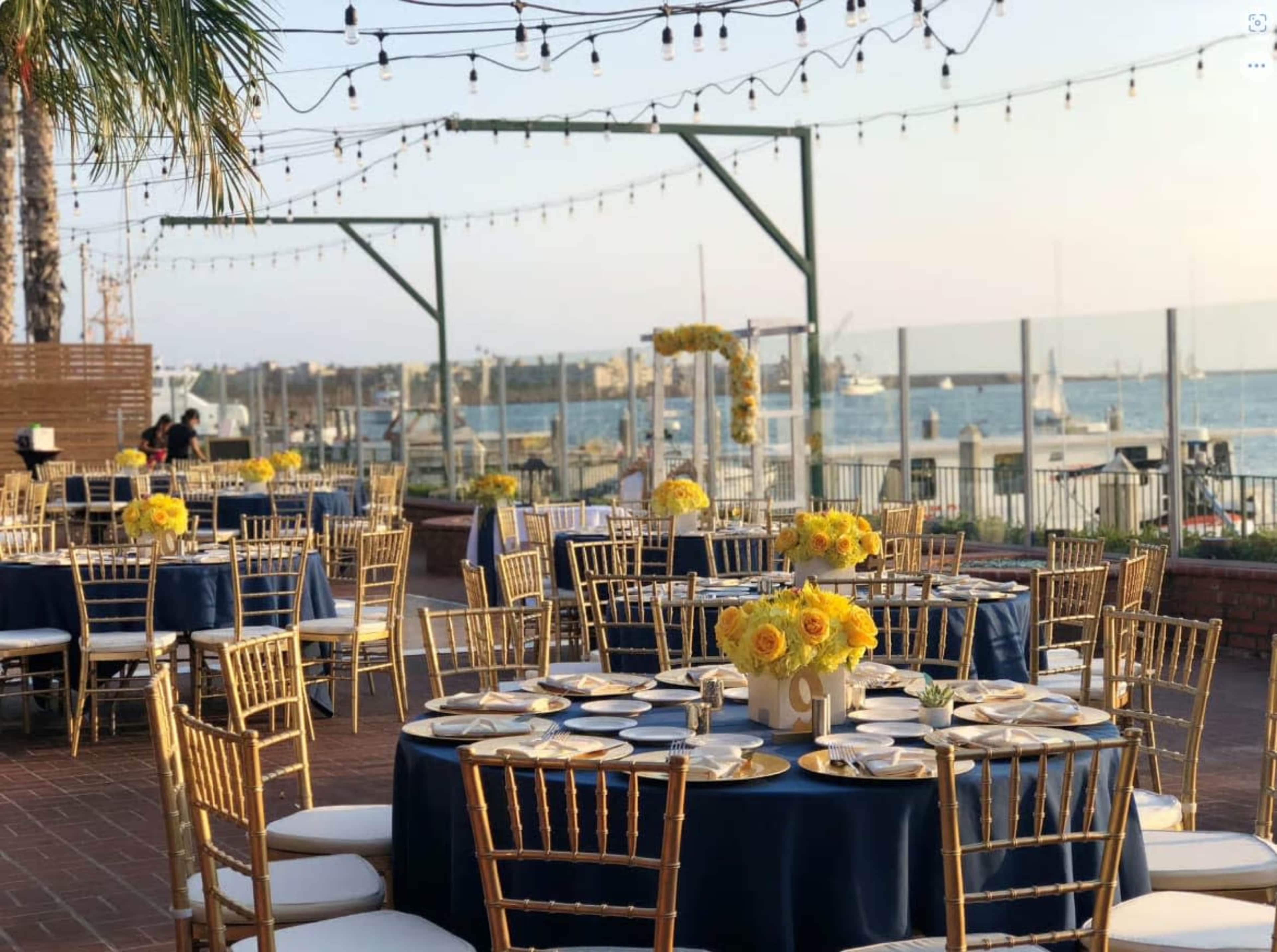 The outdoor dining area features round tables set with white tablecloths and gold chairs, adorned with yellow floral centerpieces, alongside a view of the water and string lights overhead.