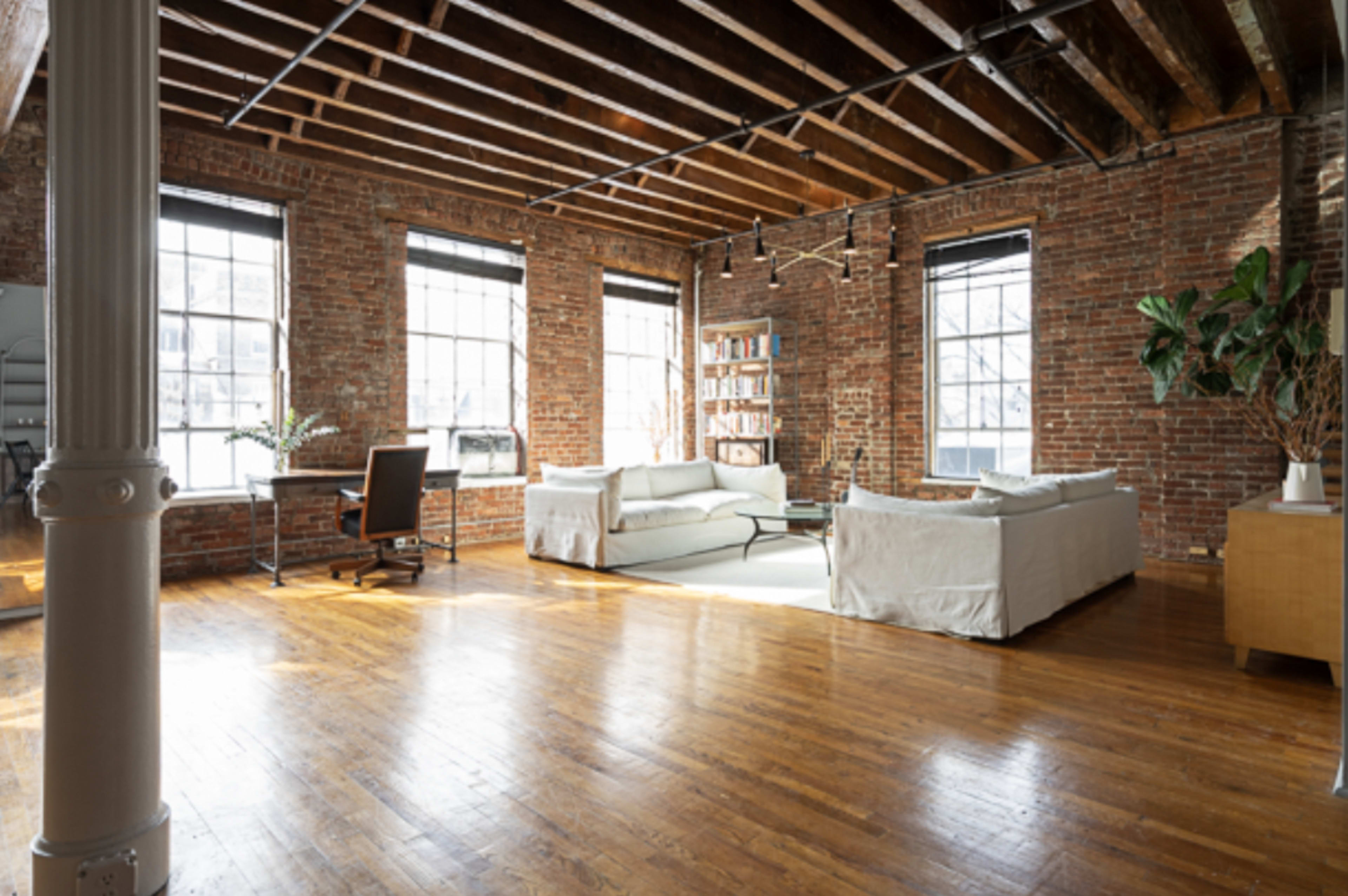 The image shows a spacious living area featuring exposed brick walls, a wooden ceiling, and hardwood flooring, with a sectional sofa, a desk, and a bookshelf.
