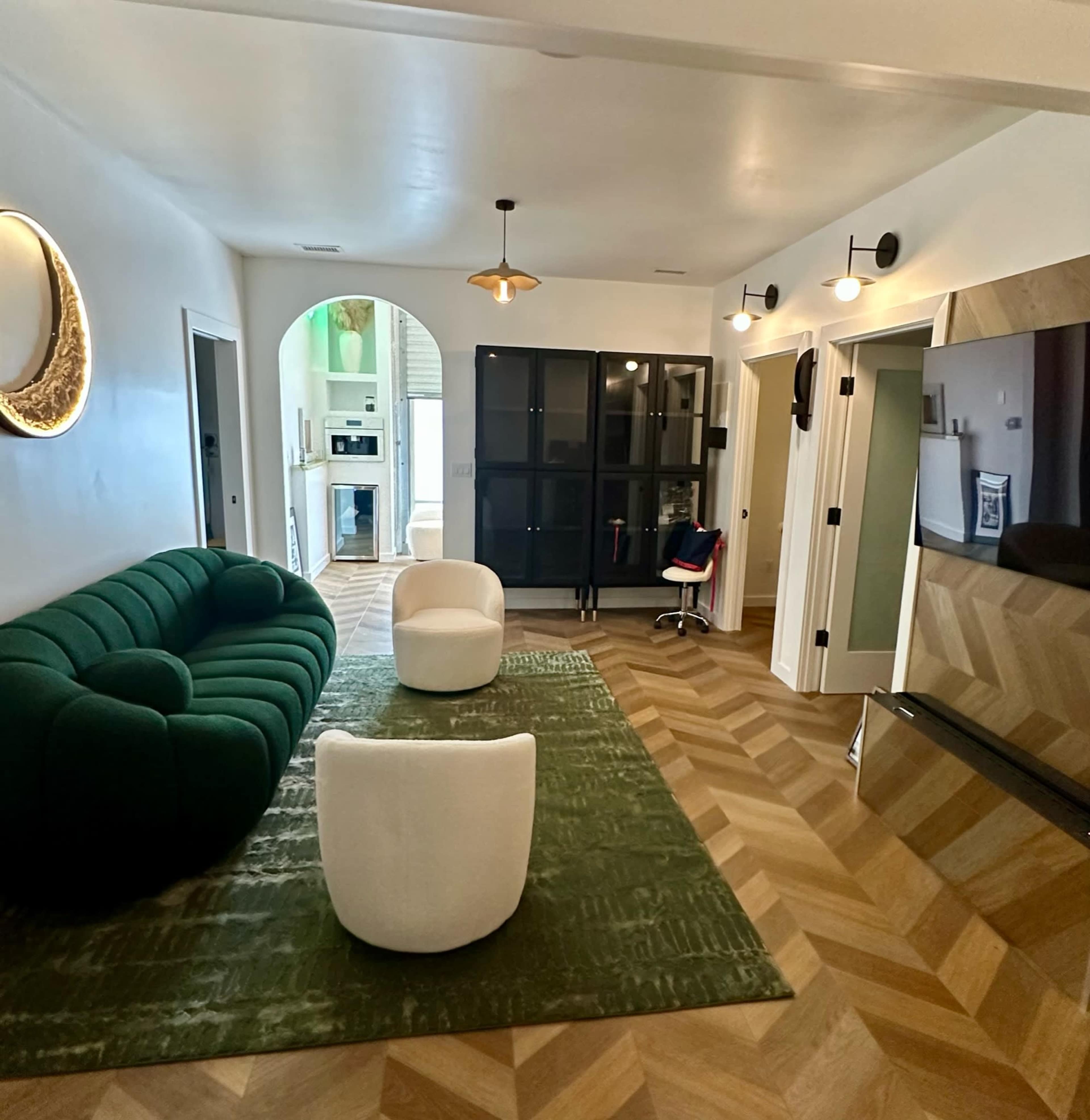A modern living room with a green tufted sofa, two white chairs, a large TV on the wall, and an arched doorway leading to a kitchen area.