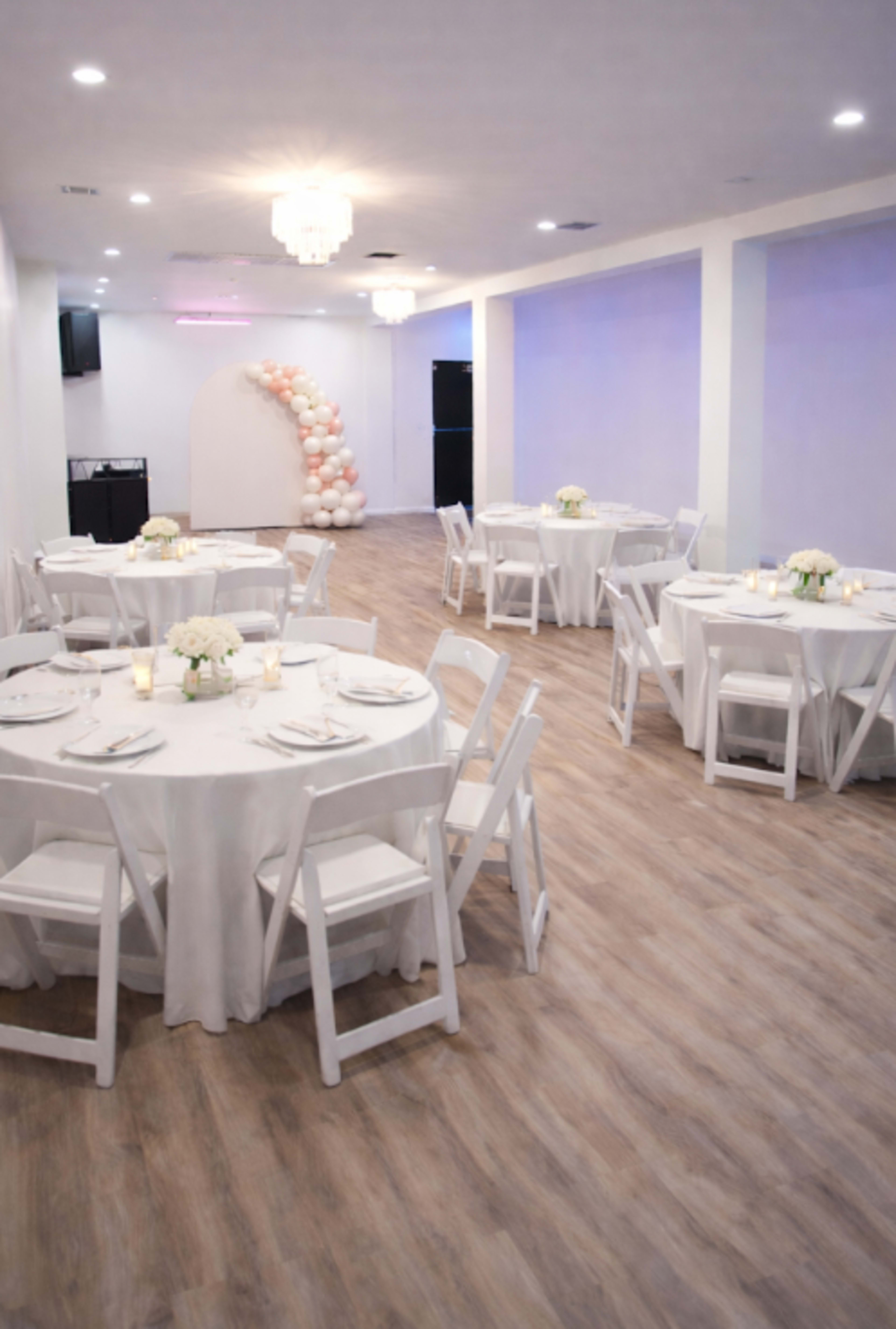 The image shows a banquet hall set up with round tables covered in white tablecloths and floral centerpieces, with a decorative balloon arch in the background.