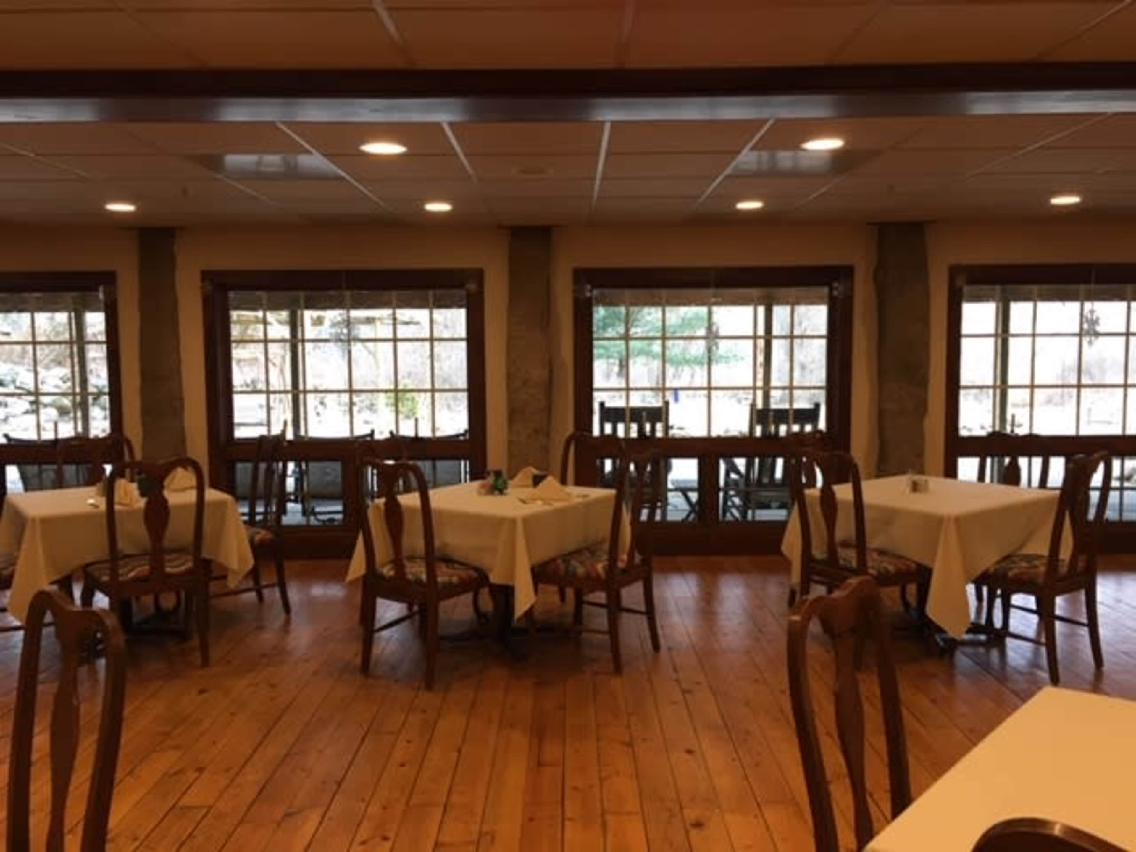 A rustic dining area with several empty tables and large windows revealing a view of trees outside.
