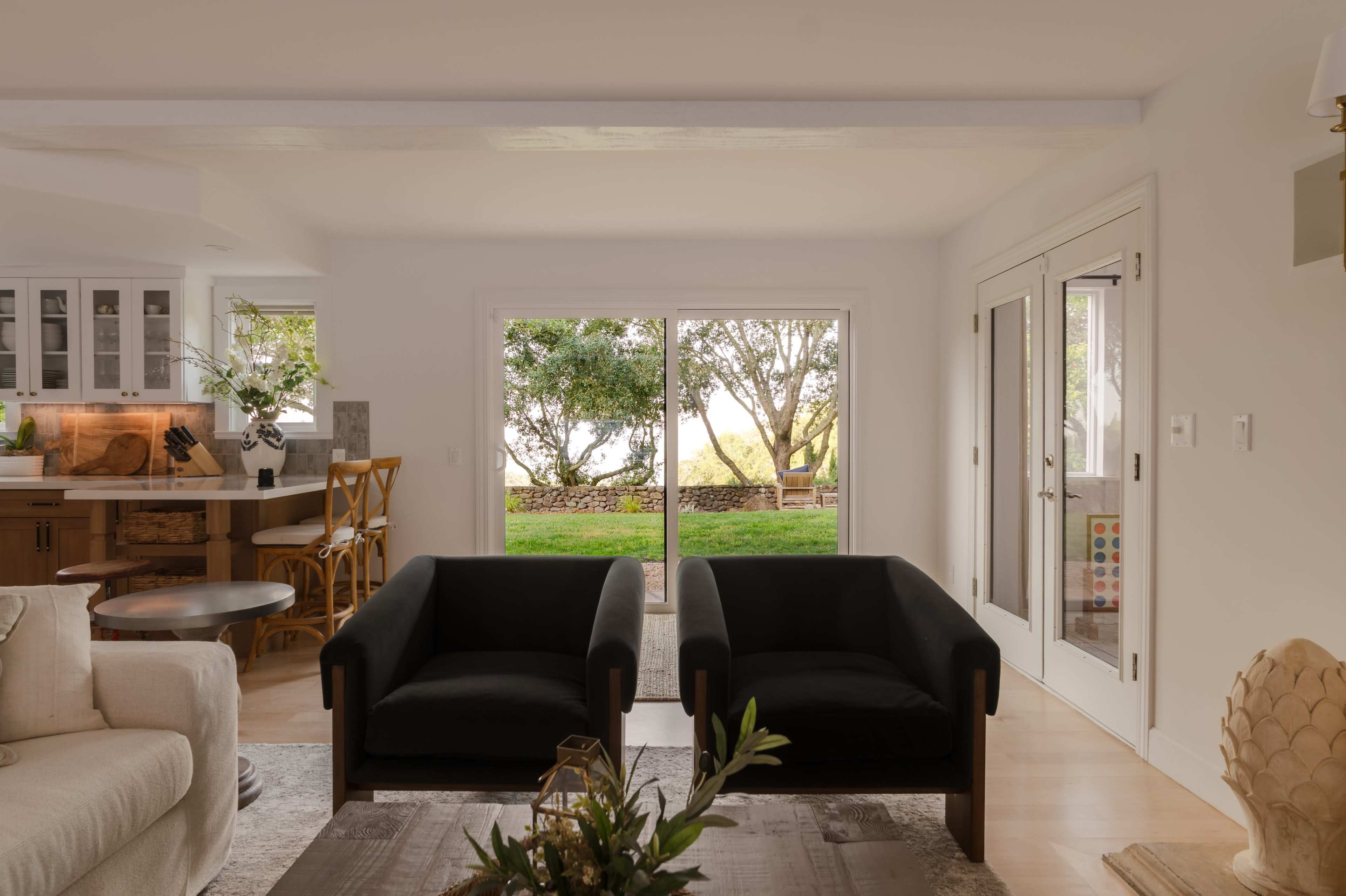The image shows a modern living room with two black chairs facing large glass doors that open to a green outdoor space.
