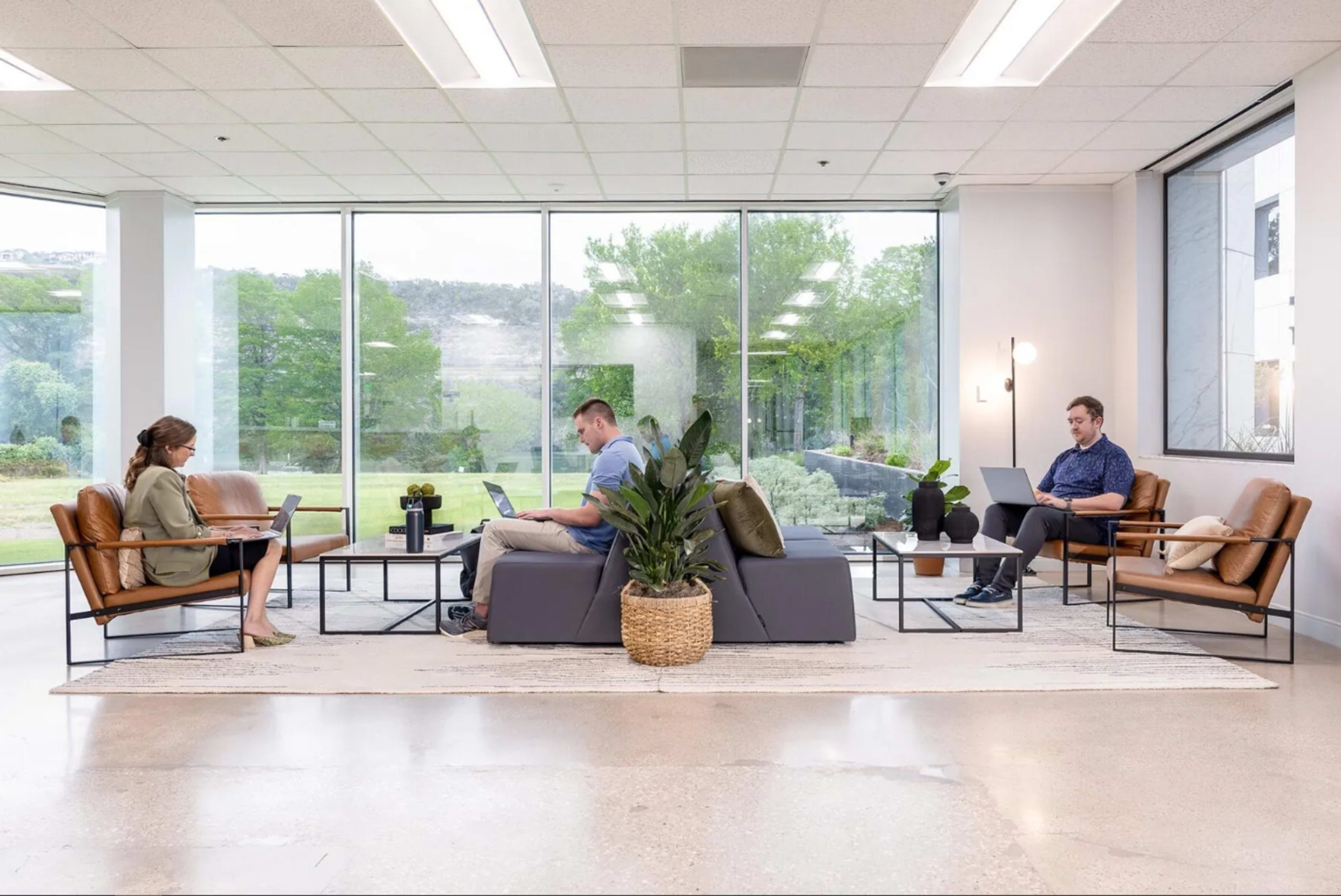 A modern office lounge features three individuals engaged in work, with a large window overlooking greenery and a comfortable seating area.