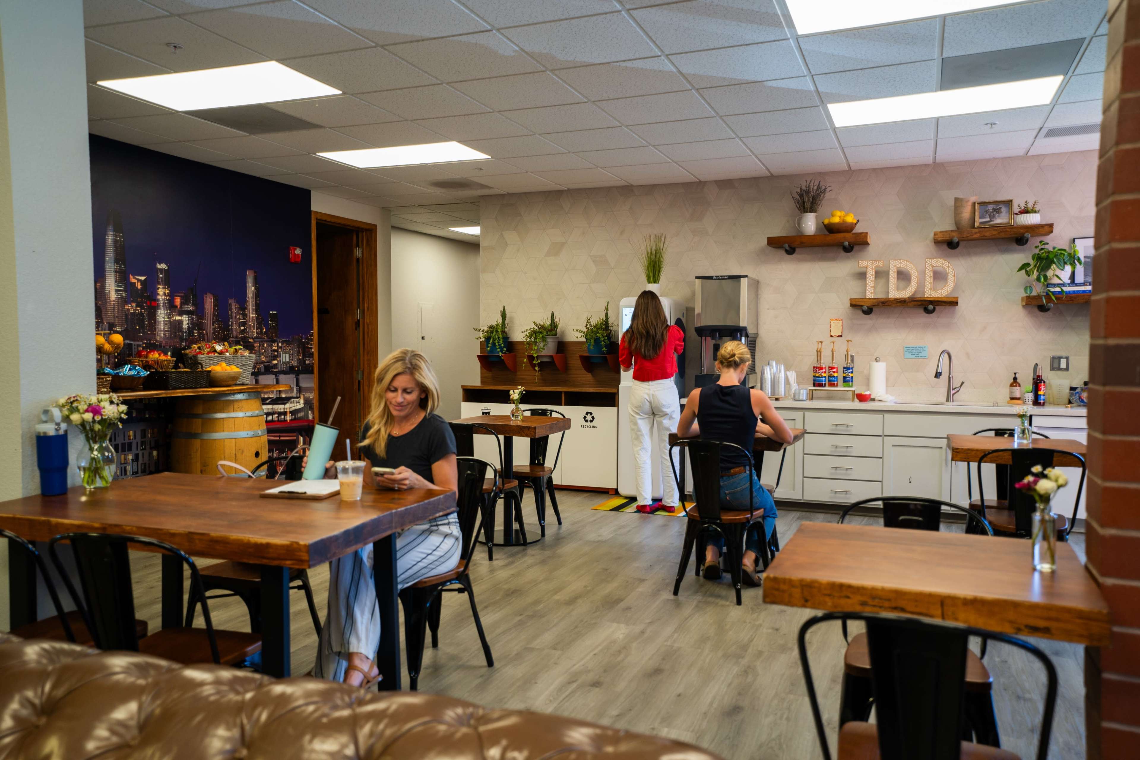 The image shows a modern office break room with two women engaged in different activities, surrounded by wooden tables and plants.