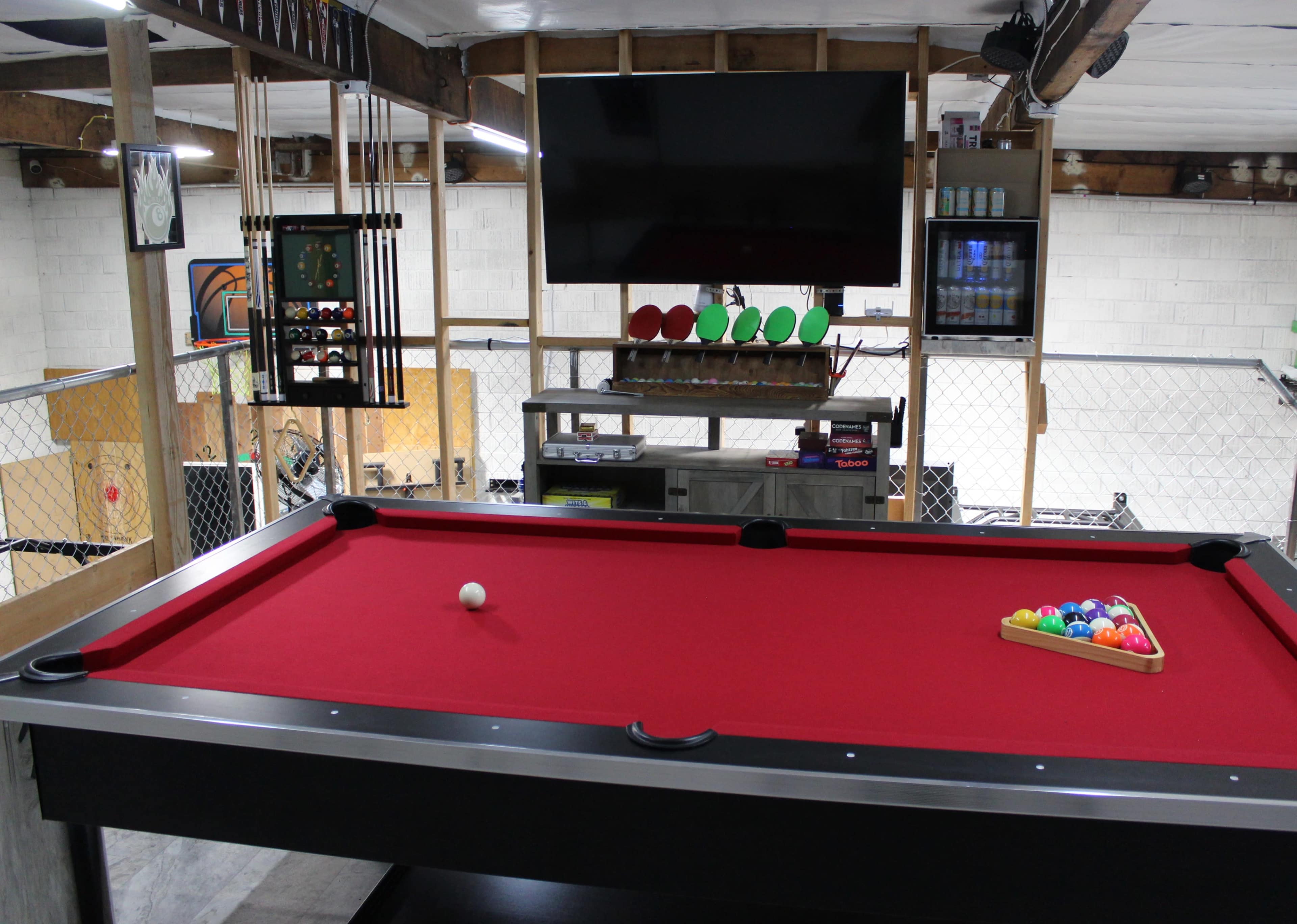 The image shows a game room featuring a red pool table, a television mounted on the wall, and a rack of pool cues.