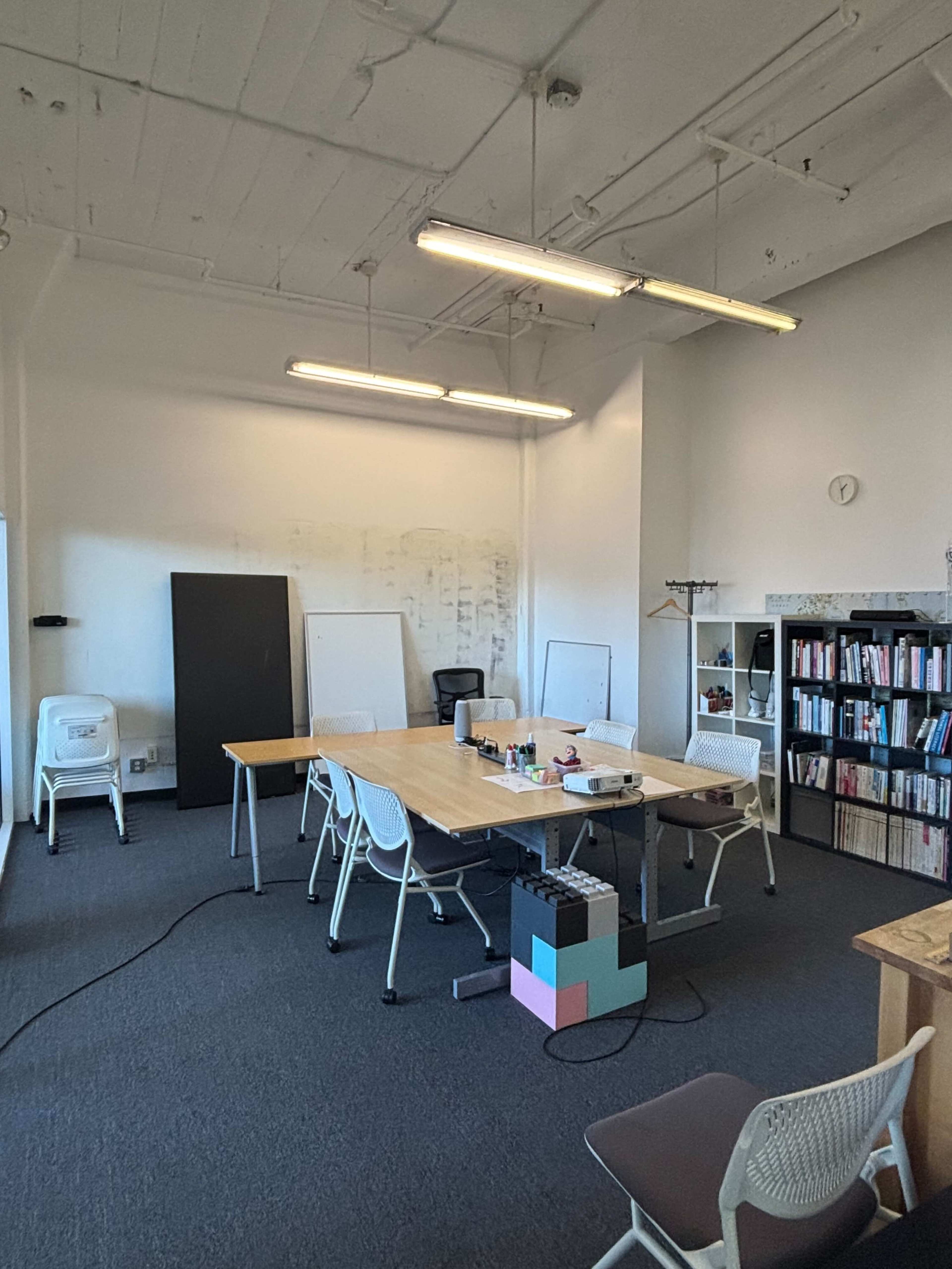 An empty office space features tables and chairs arranged for a meeting, with a bookshelf and light fixtures overhead.