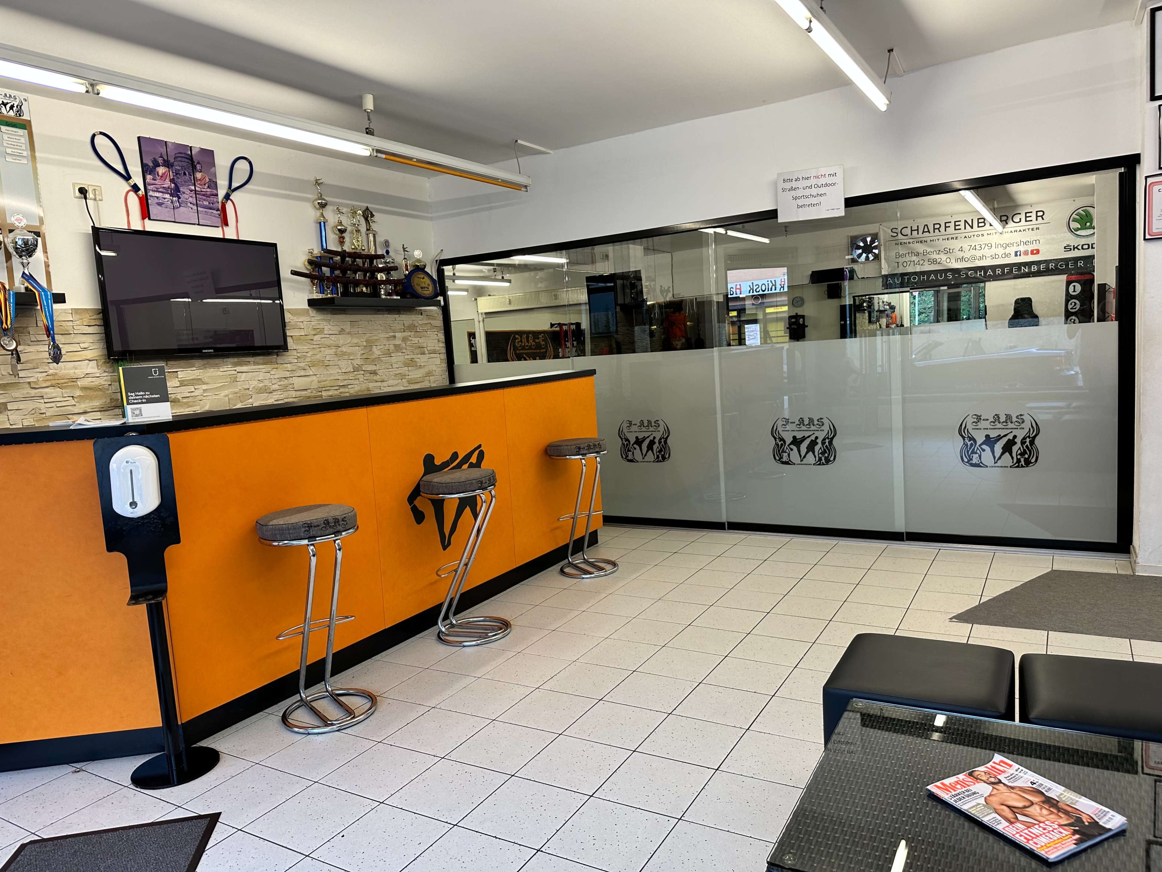 The image shows a sleek interior of a service area with an orange and white color scheme, featuring bar stools, a television, decorative trophies, and a glass partition.