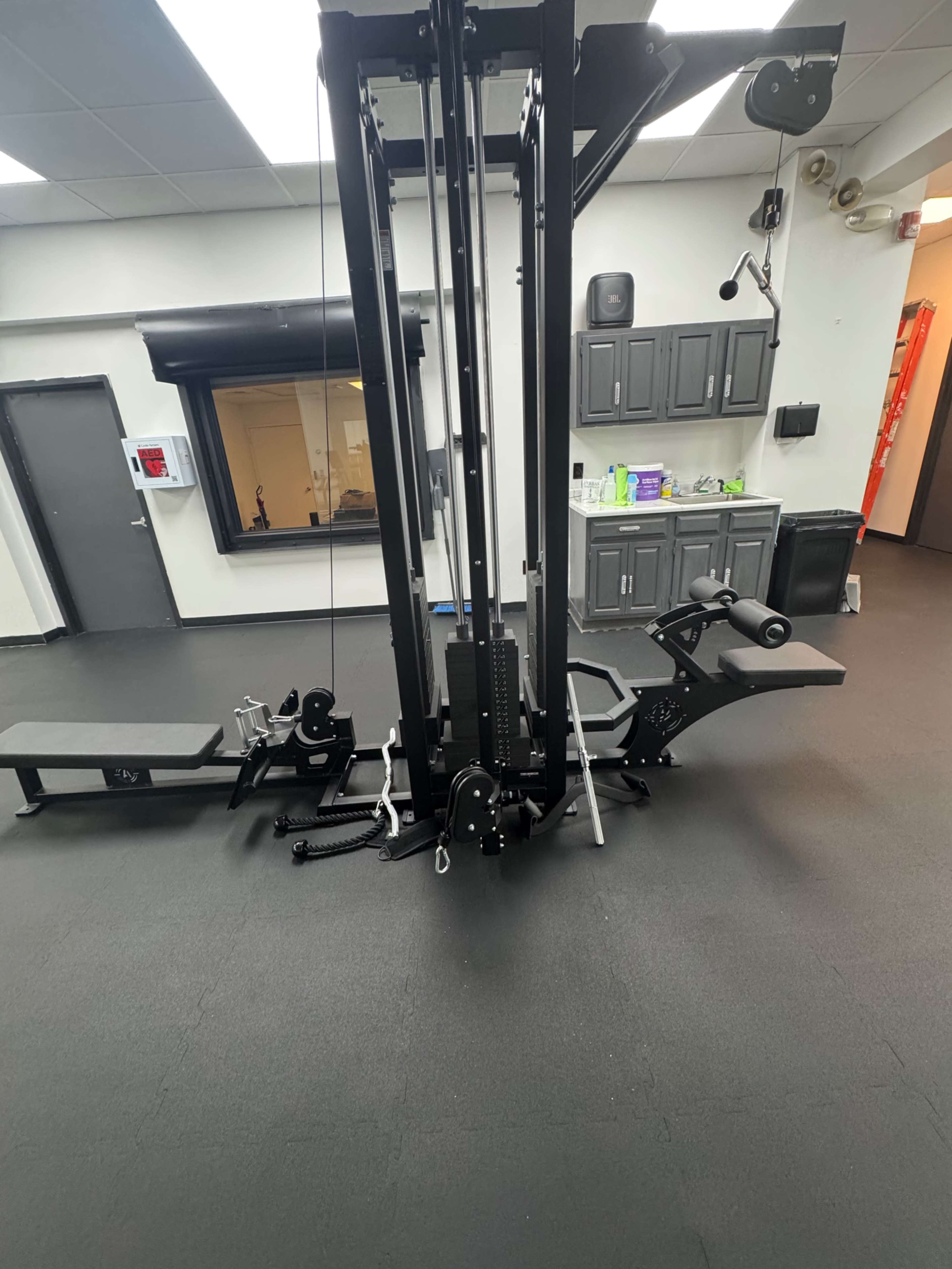 The image shows a gym equipment setup featuring a multi-station cable machine and a bench in a well-lit exercise room with gray floors and dark cabinets.