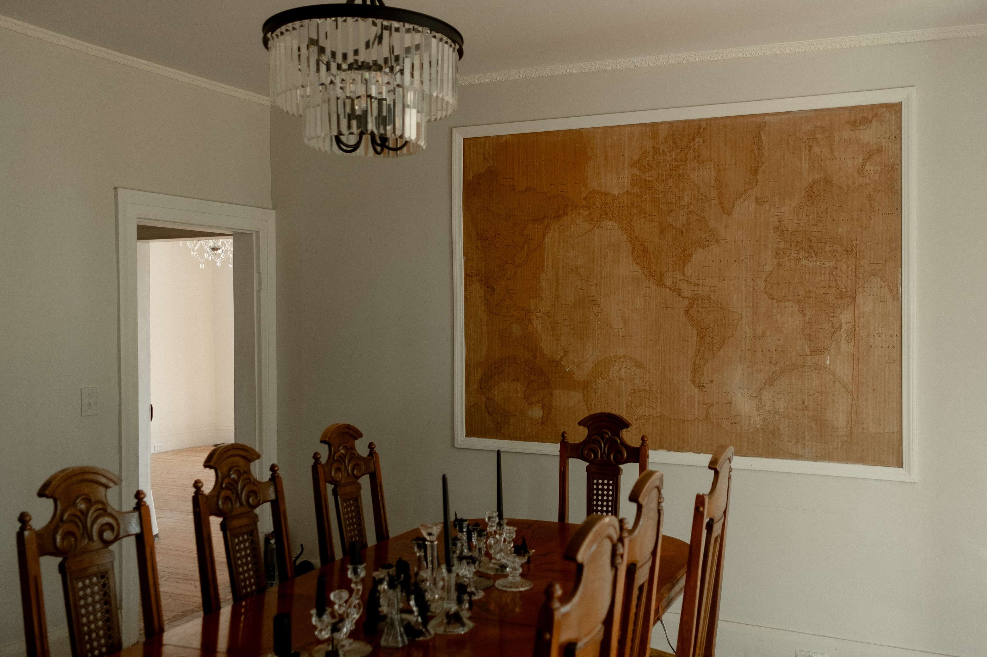 A dining room features a large wooden table set for a meal, surrounded by ornate chairs, with a world map mounted on the wall and a chandelier overhead.