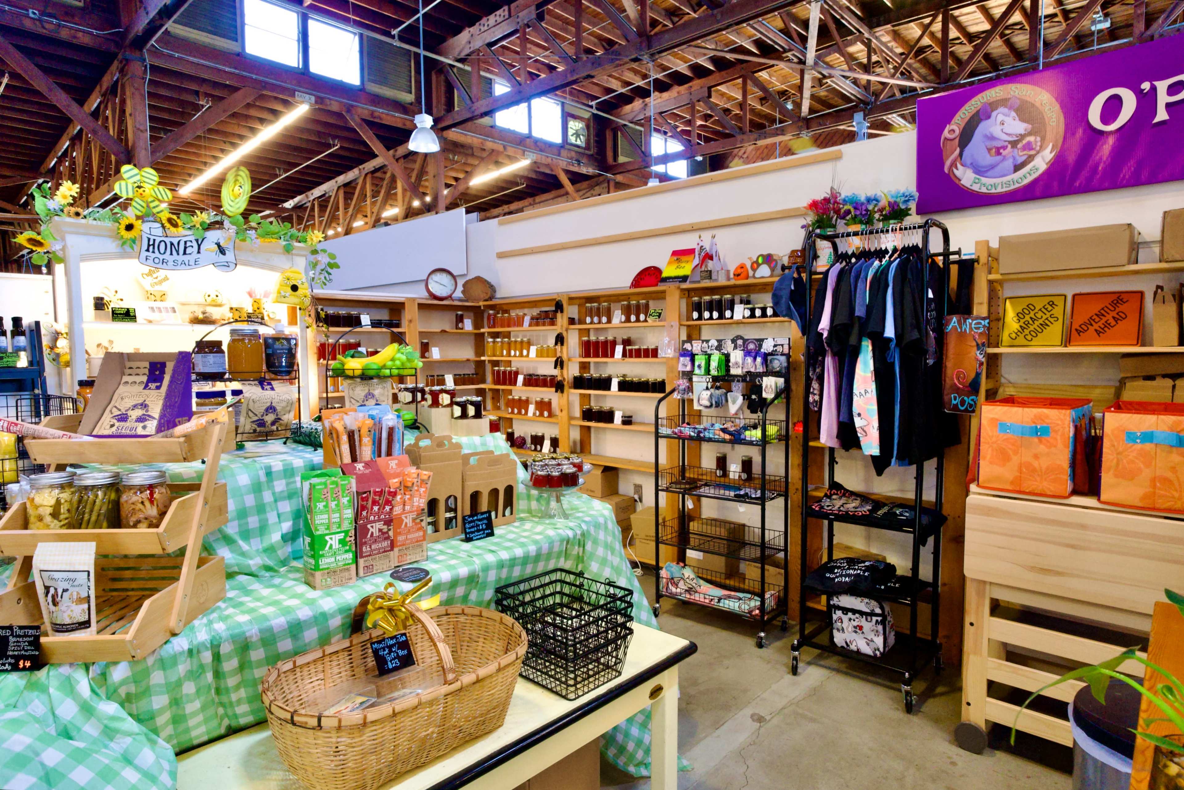 The image shows a vibrant market space with wooden beams, featuring shelves stocked with jars, packaged goods, and various products, while a checkered tablecloth displays baskets and items for sale.