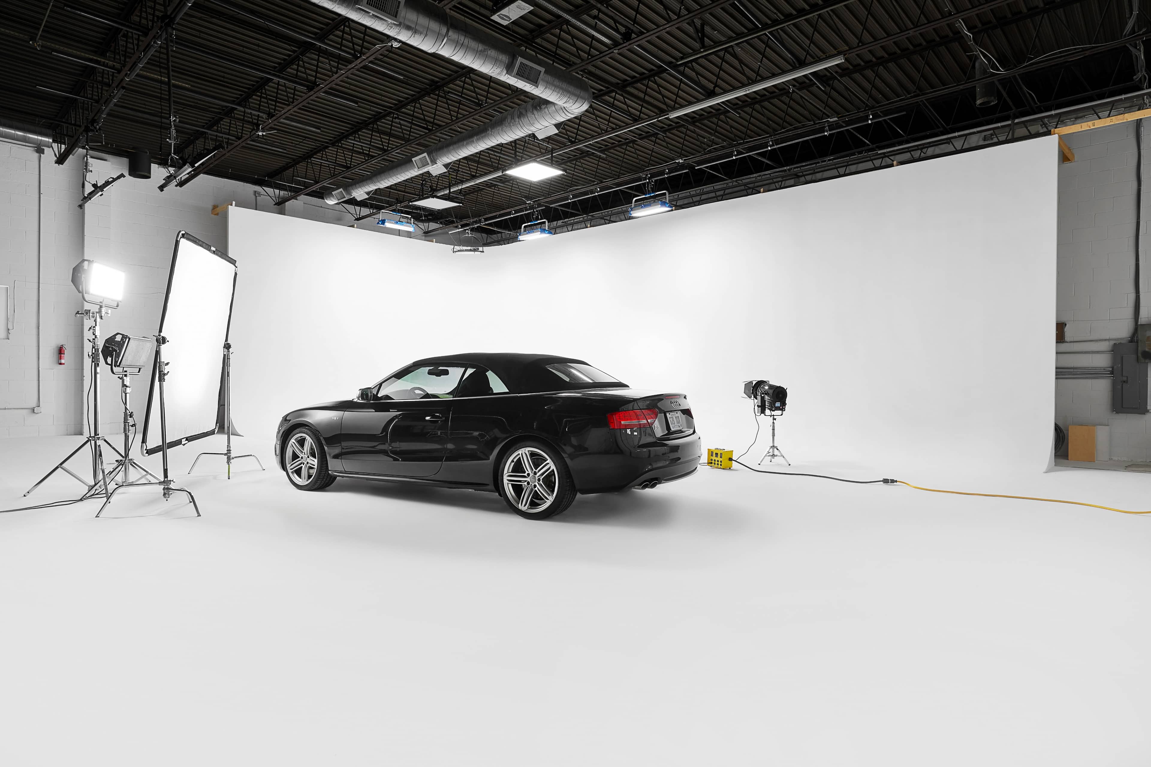 A black car is positioned in a studio with a white backdrop and professional lighting equipment.