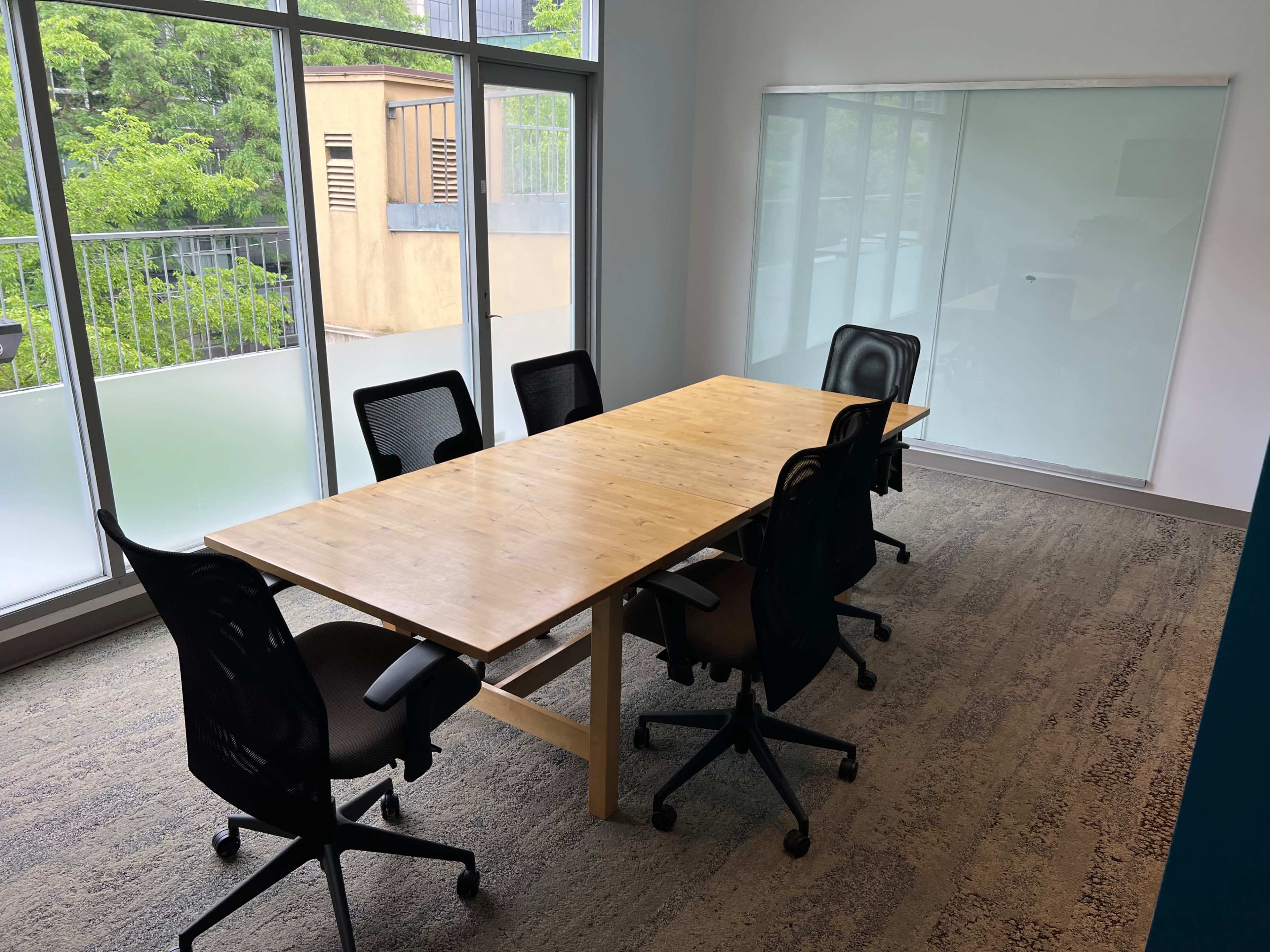 A wooden conference table surrounded by six black office chairs is located in a bright room with large windows overlooking greenery.