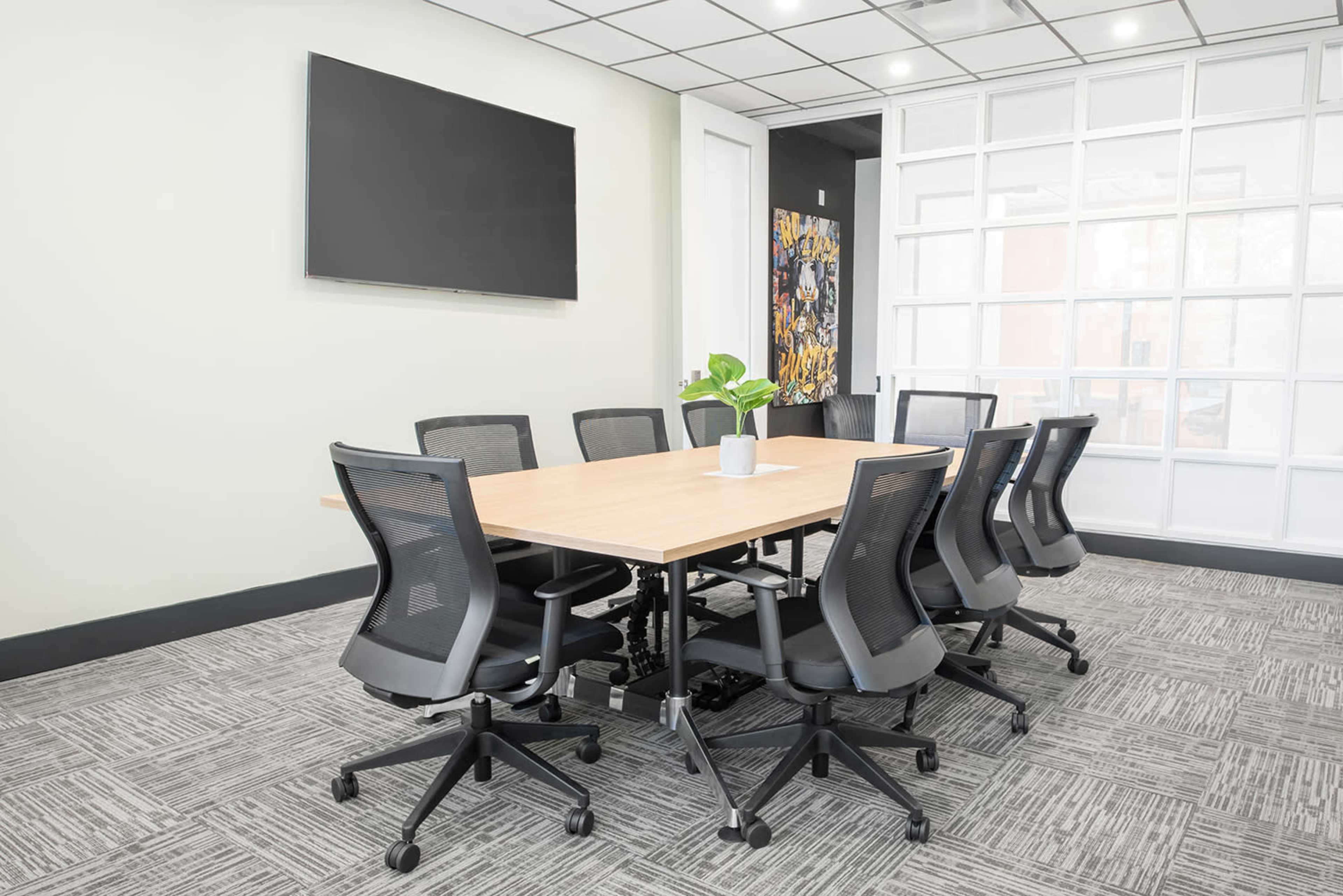 The image shows a modern conference room with a large wooden table surrounded by eight ergonomic chairs, a wall-mounted TV, and a potted plant in the center.