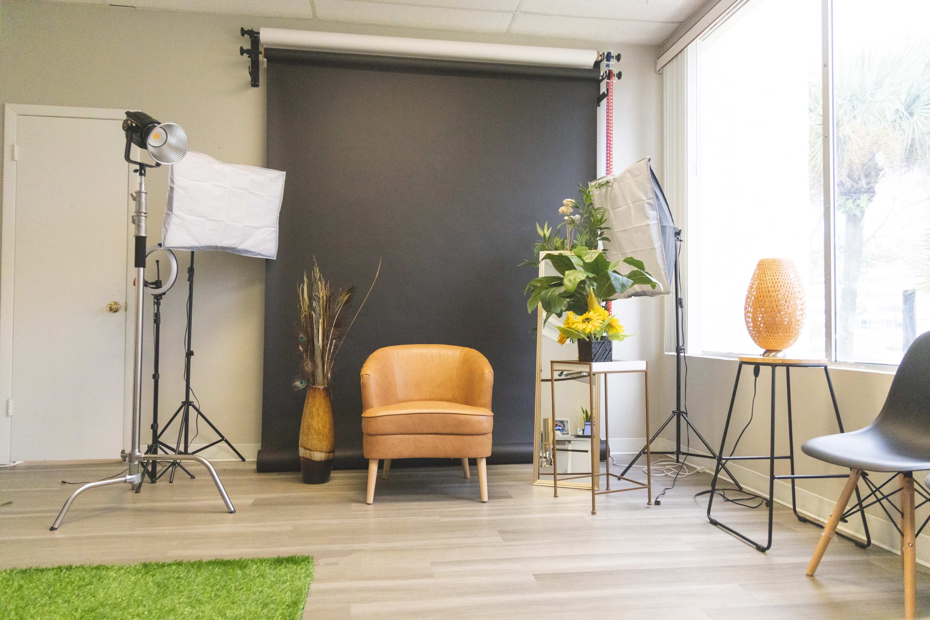 A photography studio setup featuring a brown chair, a black backdrop, softbox lights, a decorative plant, and a lamp.