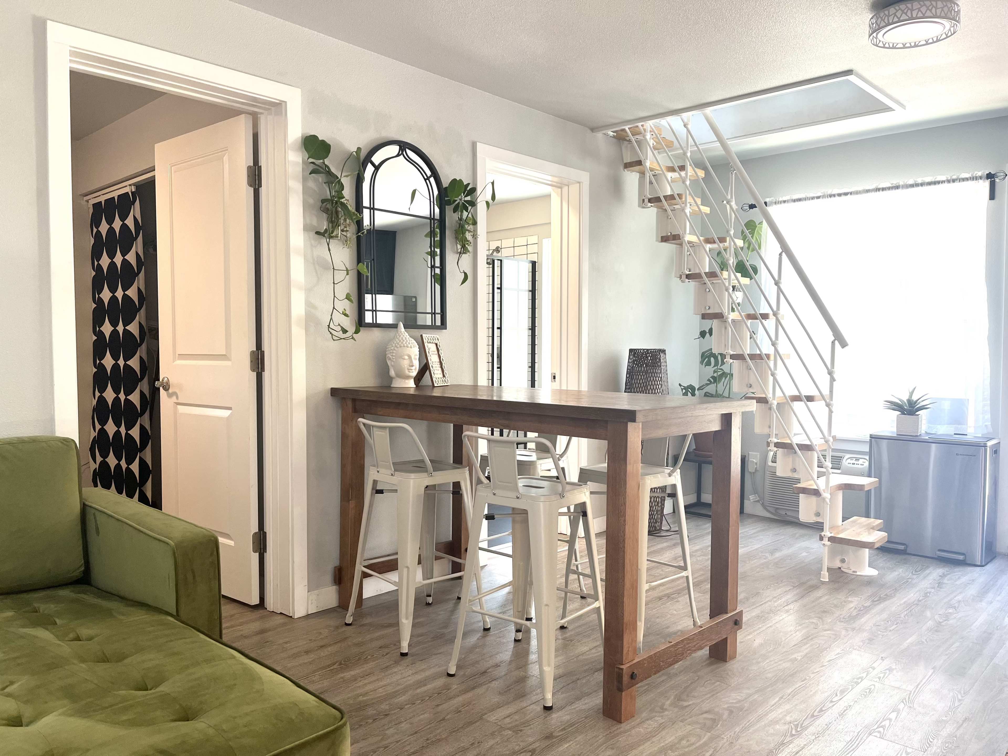 A modern living space with a green sofa, a tall wooden table with white stools, and a staircase leading to an upper level.