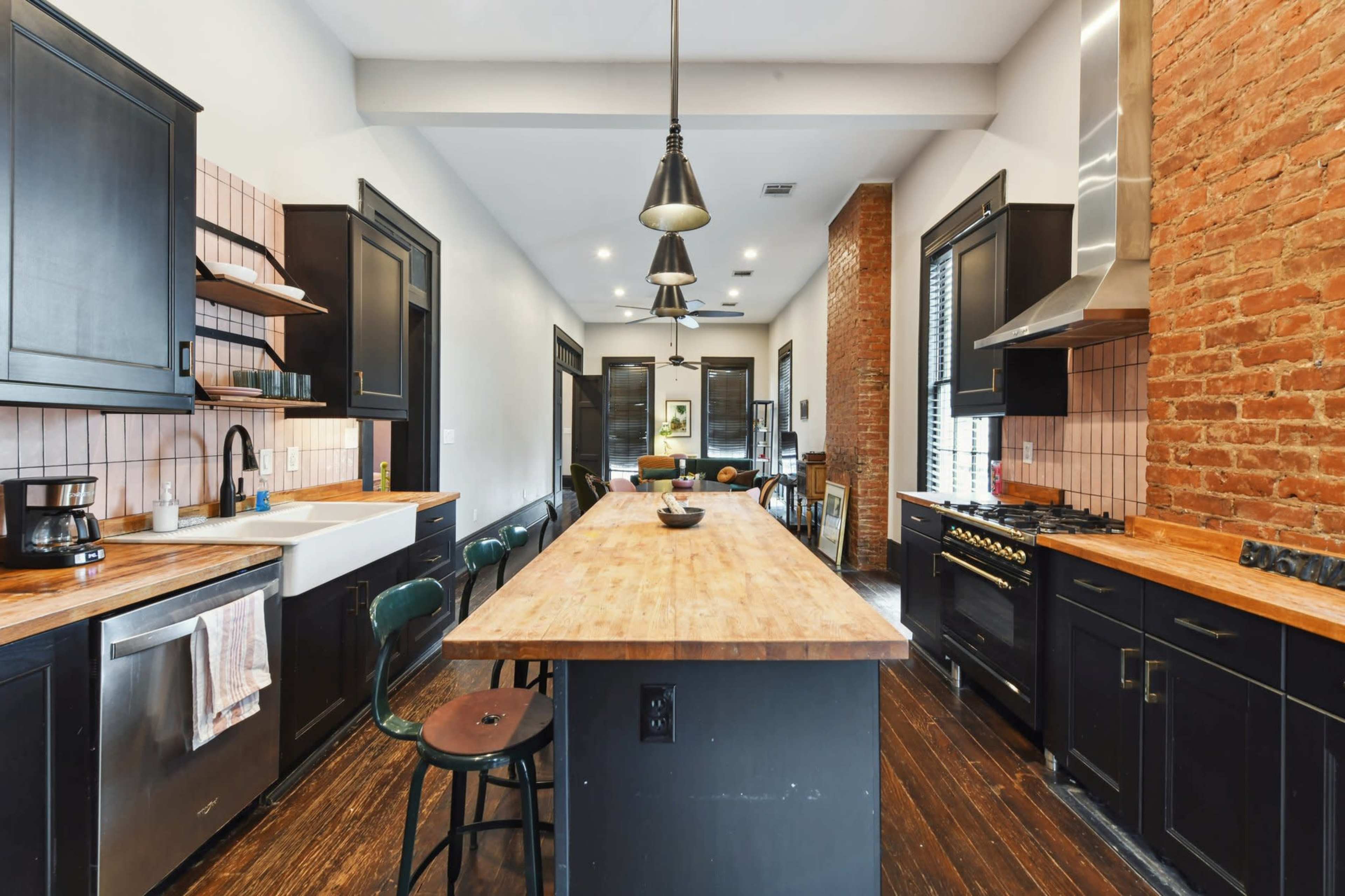 The kitchen features a long wooden island, black cabinetry, stainless steel appliances, and exposed brick walls.