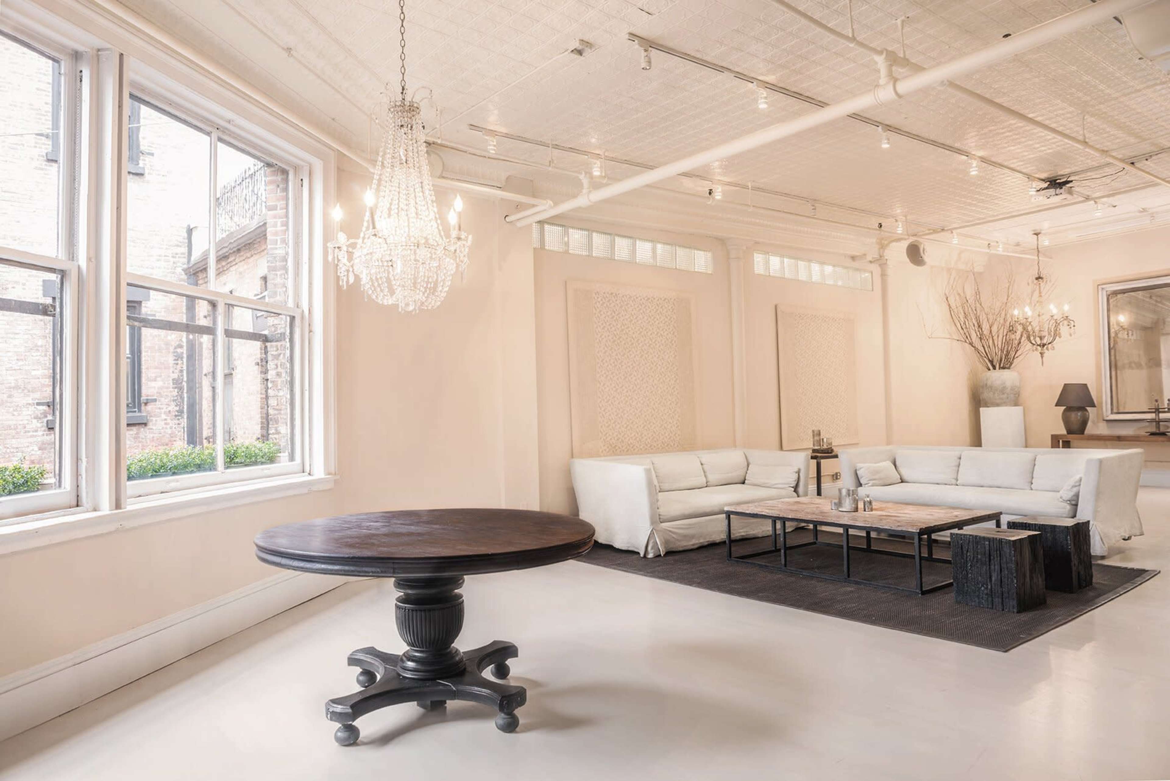 The image shows a spacious, well-lit living area featuring a round wooden table, a white sectional sofa, two coffee tables, and a chandelier hanging from the ceiling.
