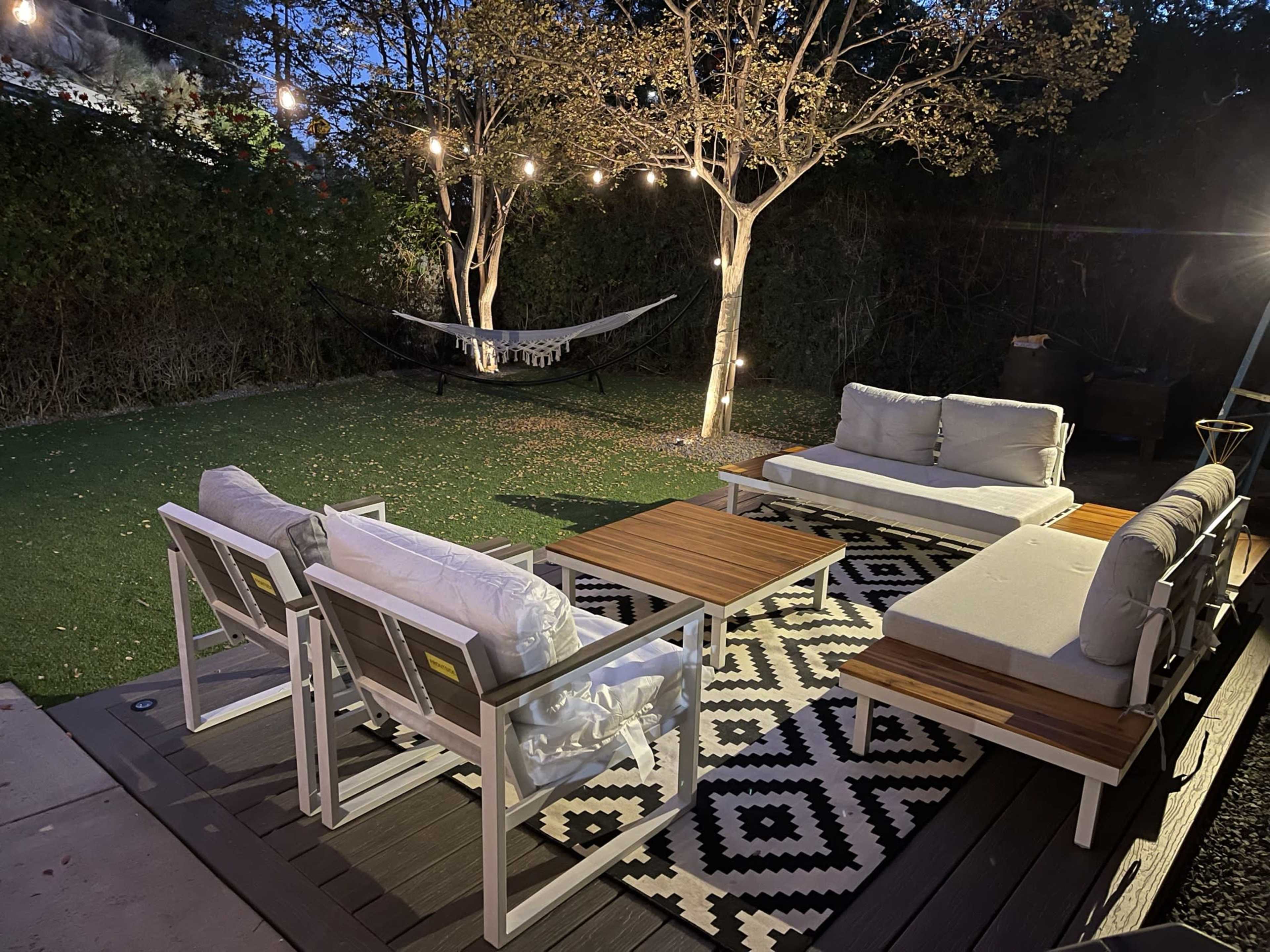 A cozy outdoor seating area features white furniture with wooden tabletops, a patterned rug, a hammock, and string lights hanging above a tree.