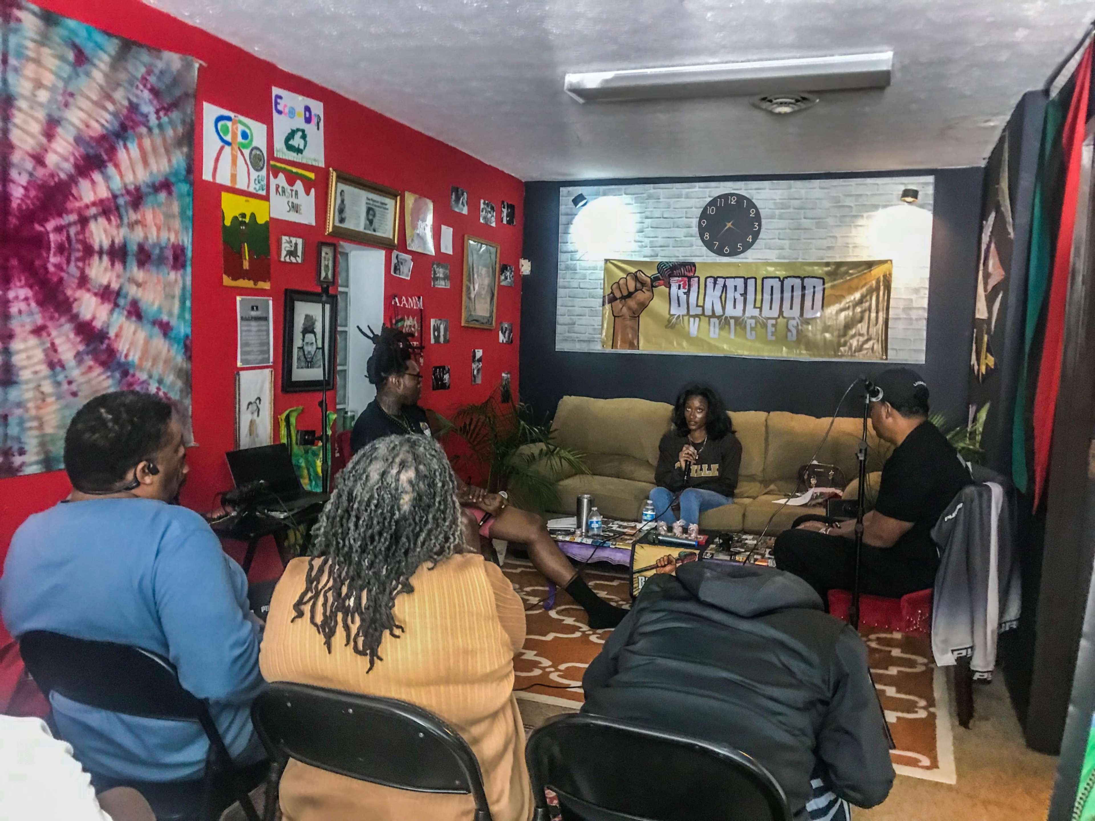 A group of people is seated in a cozy room decorated with vibrant artwork and banners, engaging in conversation around a small table.